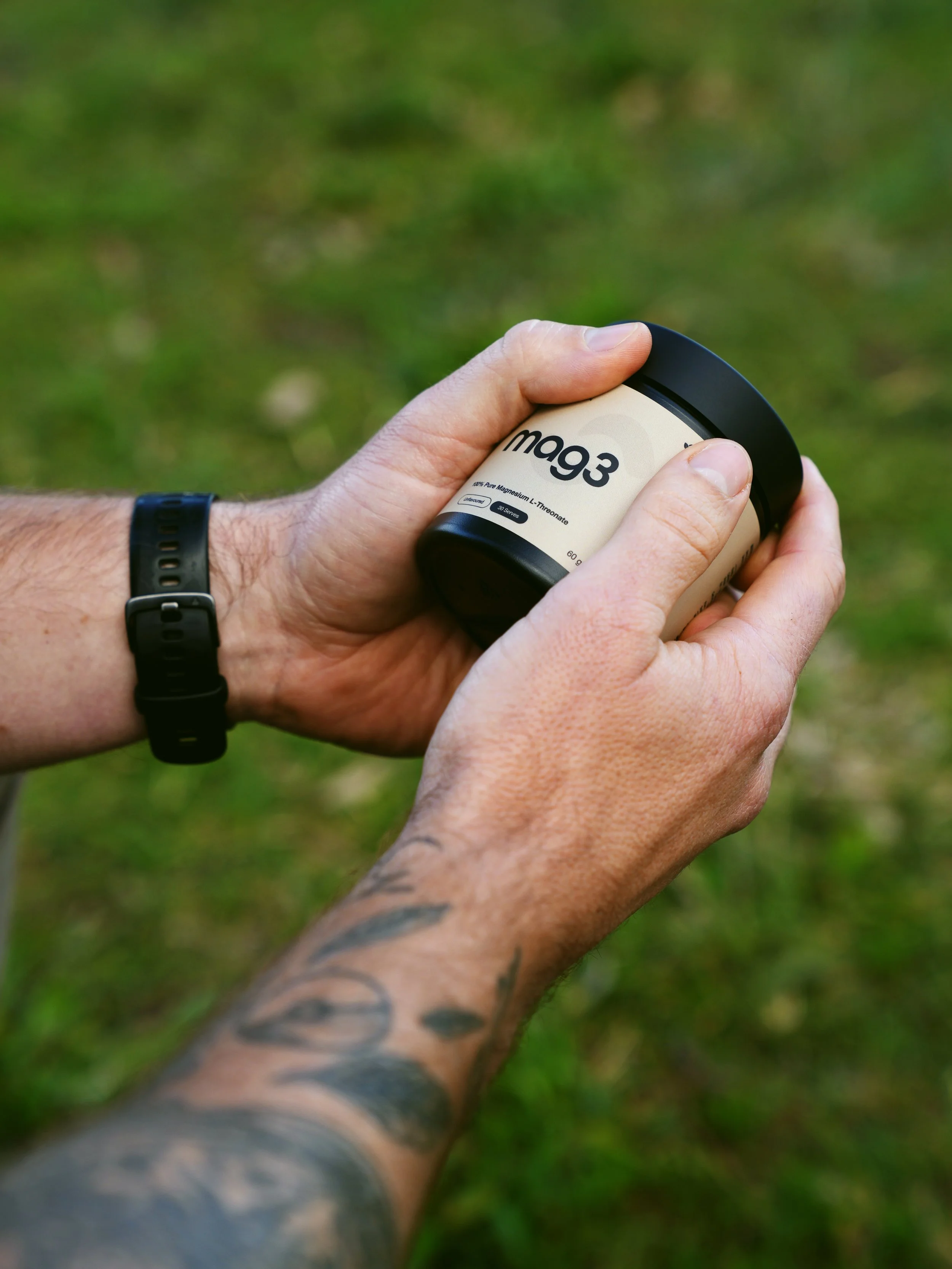 Close-up of a person's hands holding a container of magnesium supplement outdoors, with blurred green grass background.