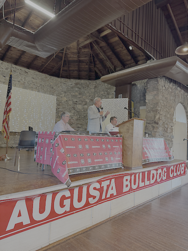 A man speaking at a podium during a meeting of the Augusta Bulldog Club, with two women seated at a table draped with Augusta Bulldog Club tablecloths, in a stone-walled room with high ceiling and ductwork.