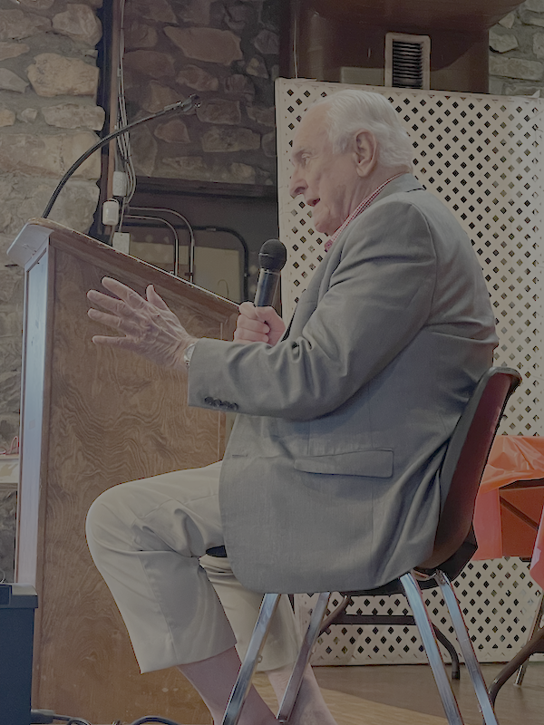 An elderly man in a gray suit and beige pants sitting on a chair, holding a microphone, and speaking or presenting at a wooden podium in a room with stone walls.