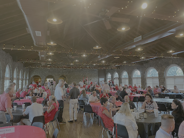 A large social event in a spacious hall with high ceilings, string lights, and large arched windows. People are sitting at round tables covered with pink and black tablecloths, engaging in conversations and dining. Some individuals are standing and serving themselves or talking.