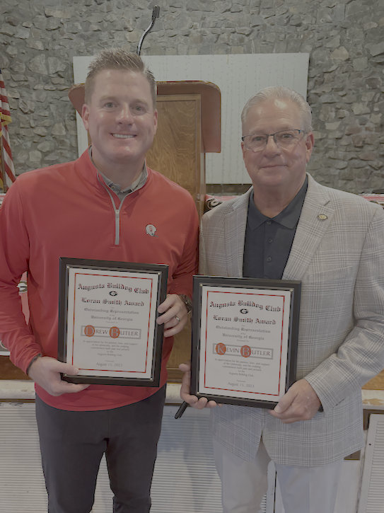 Two men standing indoors, holding awards, smiling at the camera, with a stone wall background.
