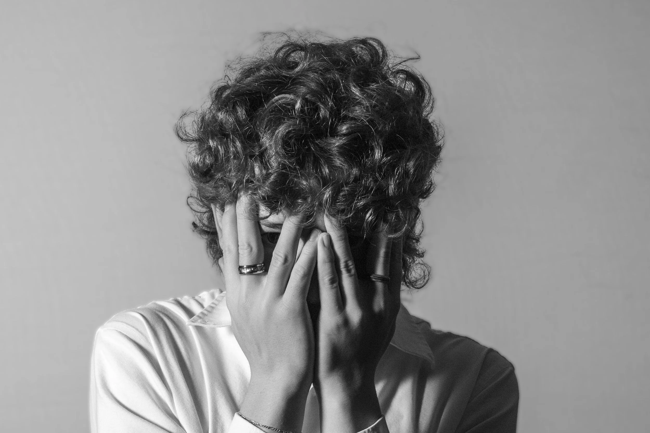 A person with curly hair covering their face with their hands, wearing rings and a bracelet, in black and white.