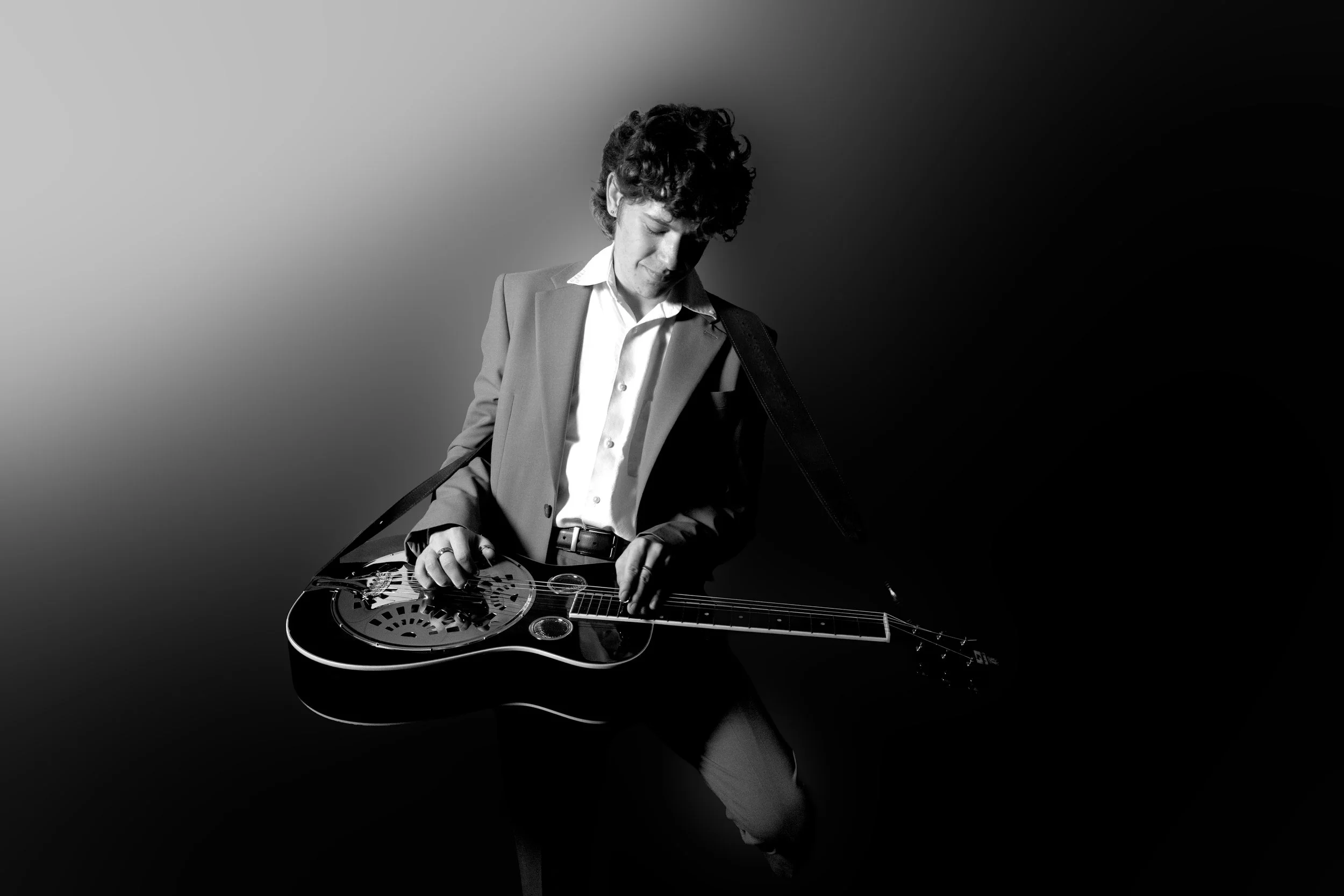 A black and white photo of a young man with curly hair wearing a suit jacket, white shirt, and black belt, playing a resonator guitar.
