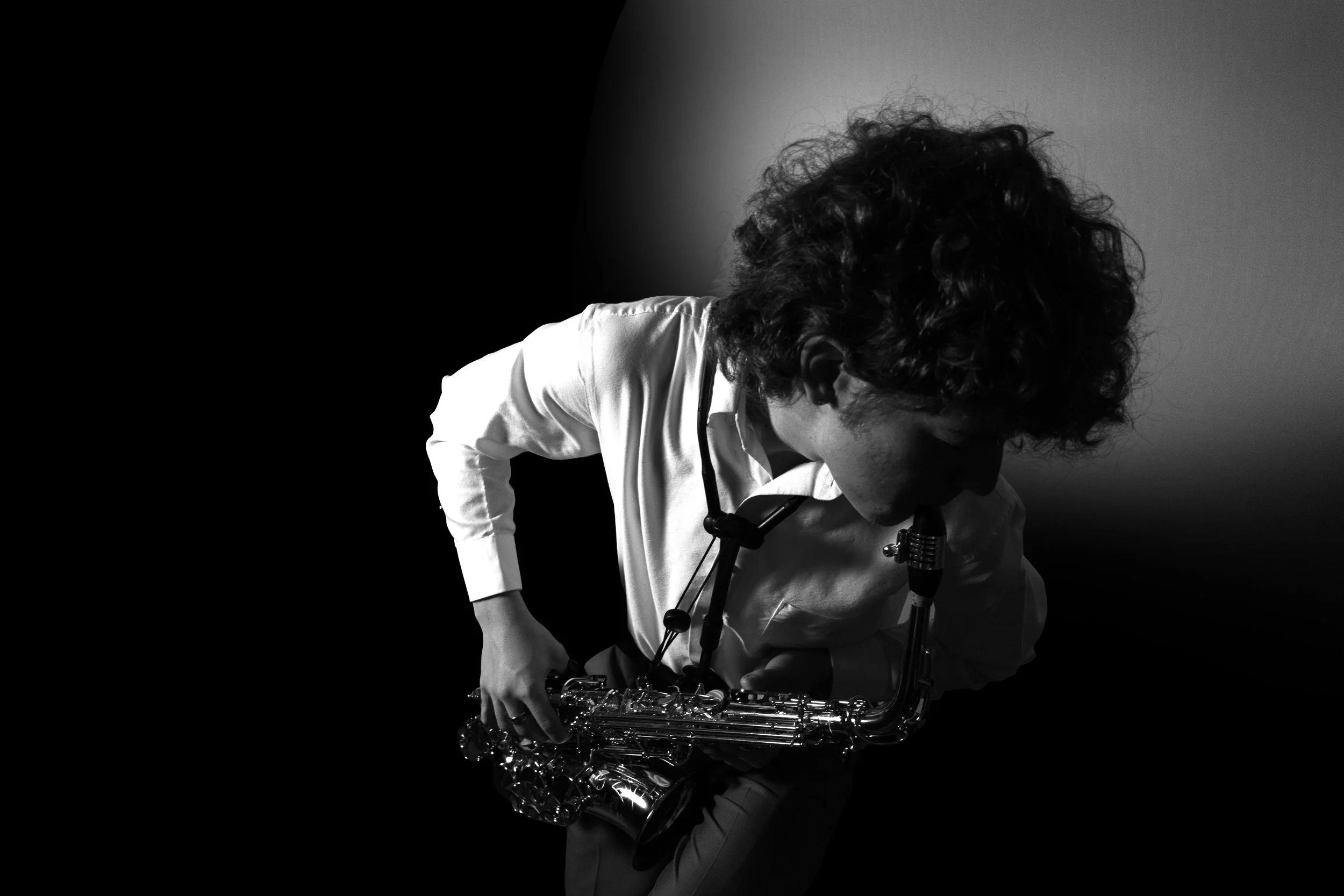 A person with curly hair wearing a white shirt and glasses, holding a saxophone, with a dark background and a gray wall.