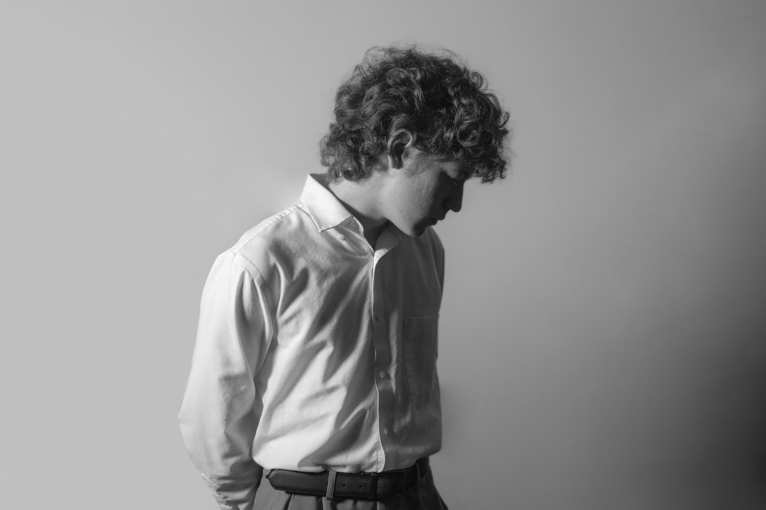 A black and white photo of a young man with curly hair, wearing a white shirt, standing against a plain wall, looking down.