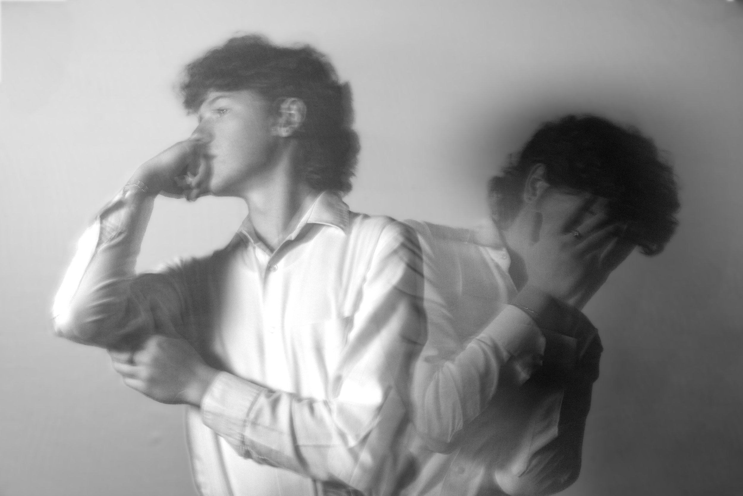 A black and white photo of a young man with curly hair, wearing glasses and a striped shirt, standing next to someone with similar hair and glasses, both with thoughtful expressions, against a plain background.