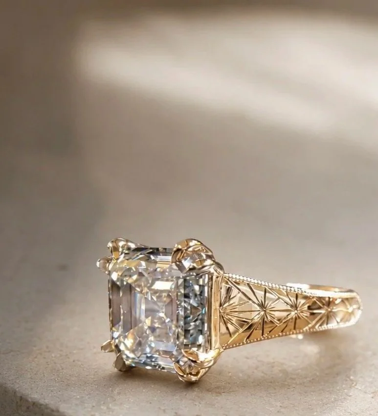 A gold engagement ring with an asscher cut diamond in a prong setting, featuring intricate engraving on the band.