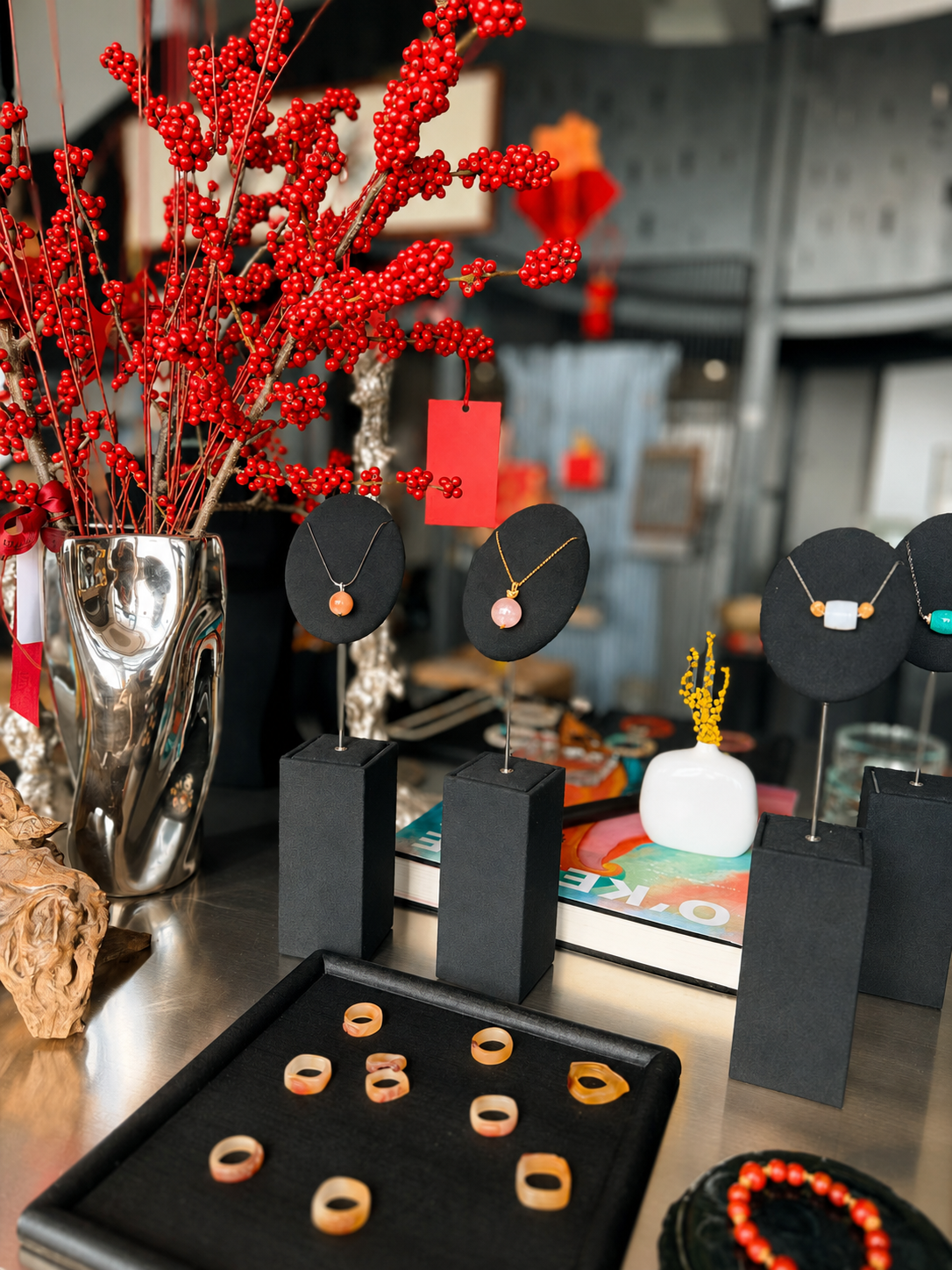 Display of jewelry including necklaces with pink and peach beads, rings on a tray, and a decorative red berry arrangement in a metallic vase, set on a table in a shop or boutique.