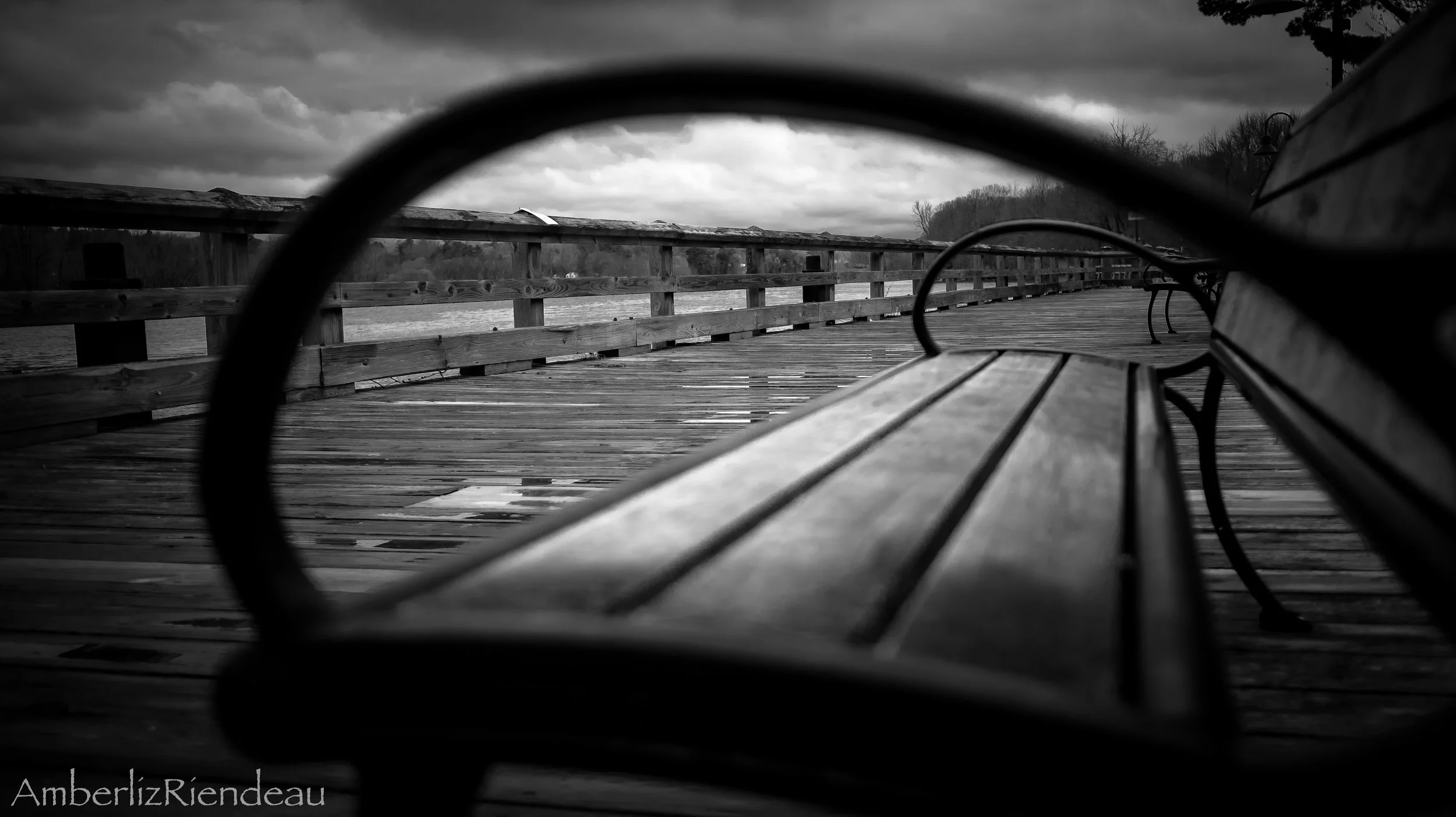 Black and white photo of a wooden bench on a weathered dock by a body of water, with cloudy sky and trees in the background, viewed through a circular frame.