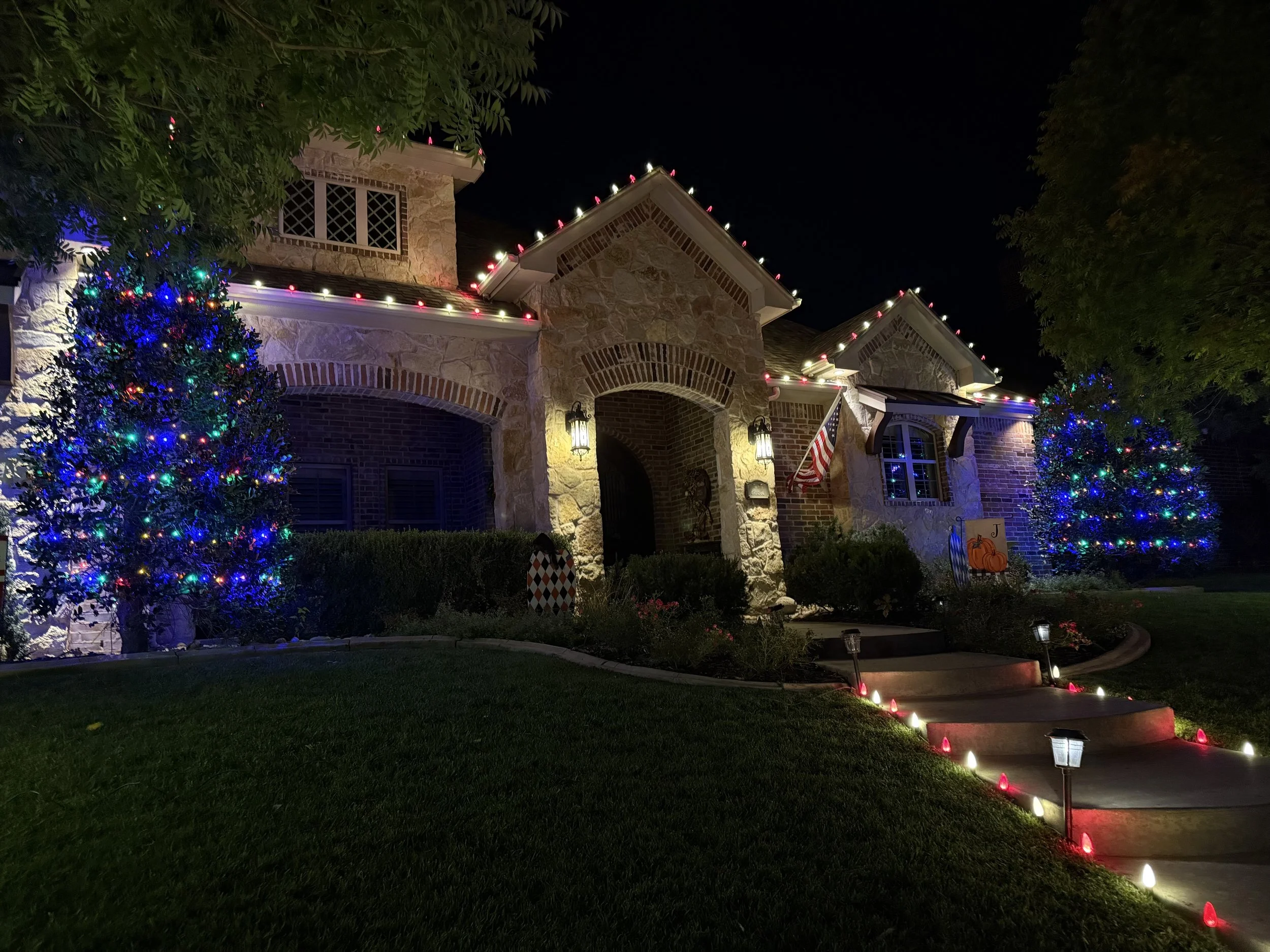 Nighttime view of a house decorated with multicolored Christmas lights along the roofline and bushes, with a lit pathway and a pumpkin decoration on the lawn.
