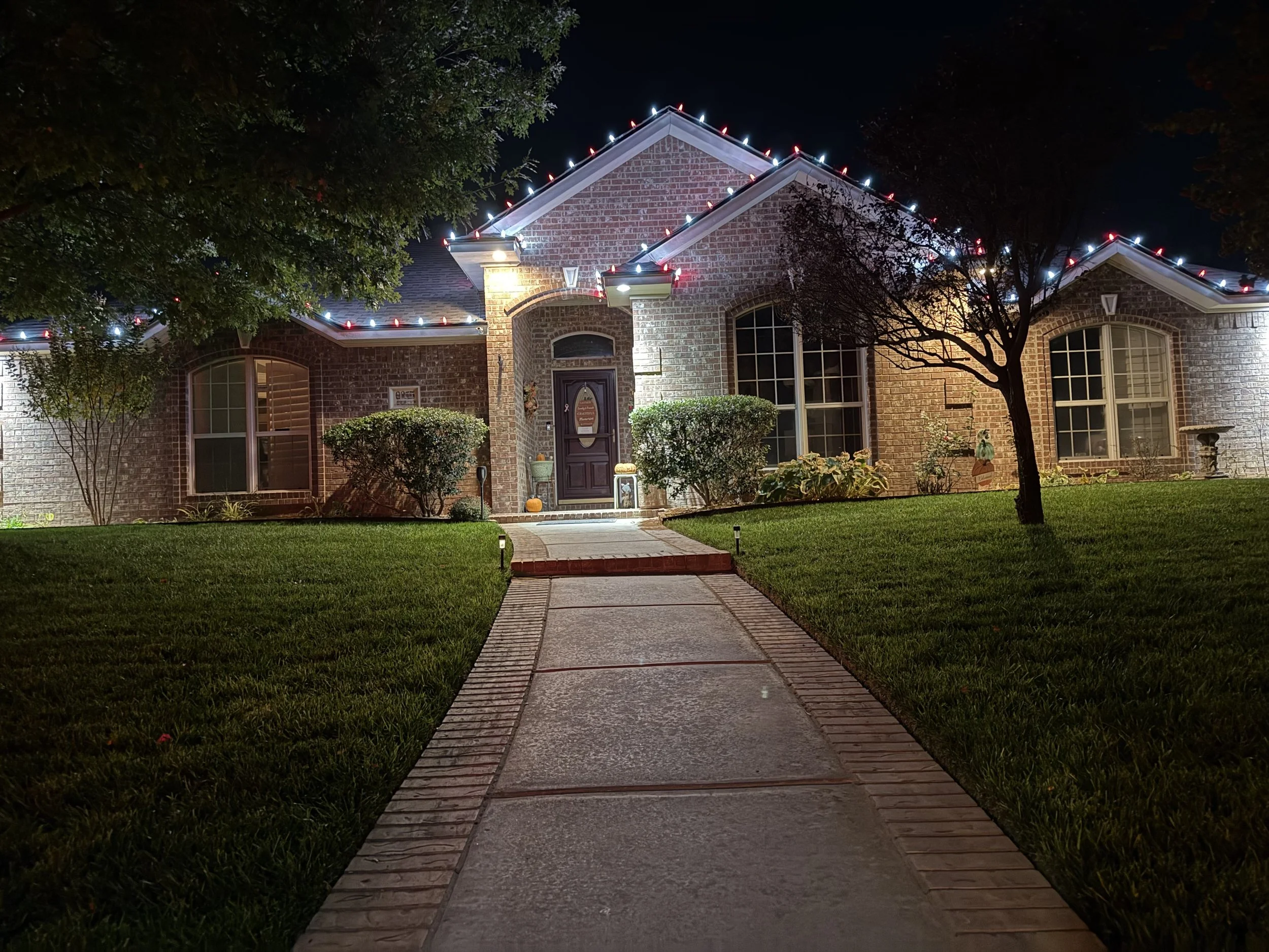 Night view of a brick house decorated with colorful Christmas lights along the roofline, surrounded by trees and a lawn, with a concrete walkway leading to the front door.