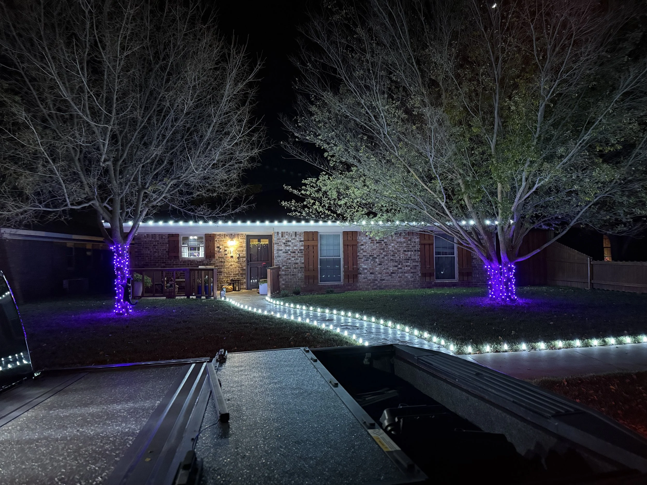 Nighttime view of a house with brick exterior, two trees decorated with purple string lights, and a pathway lined with string lights.