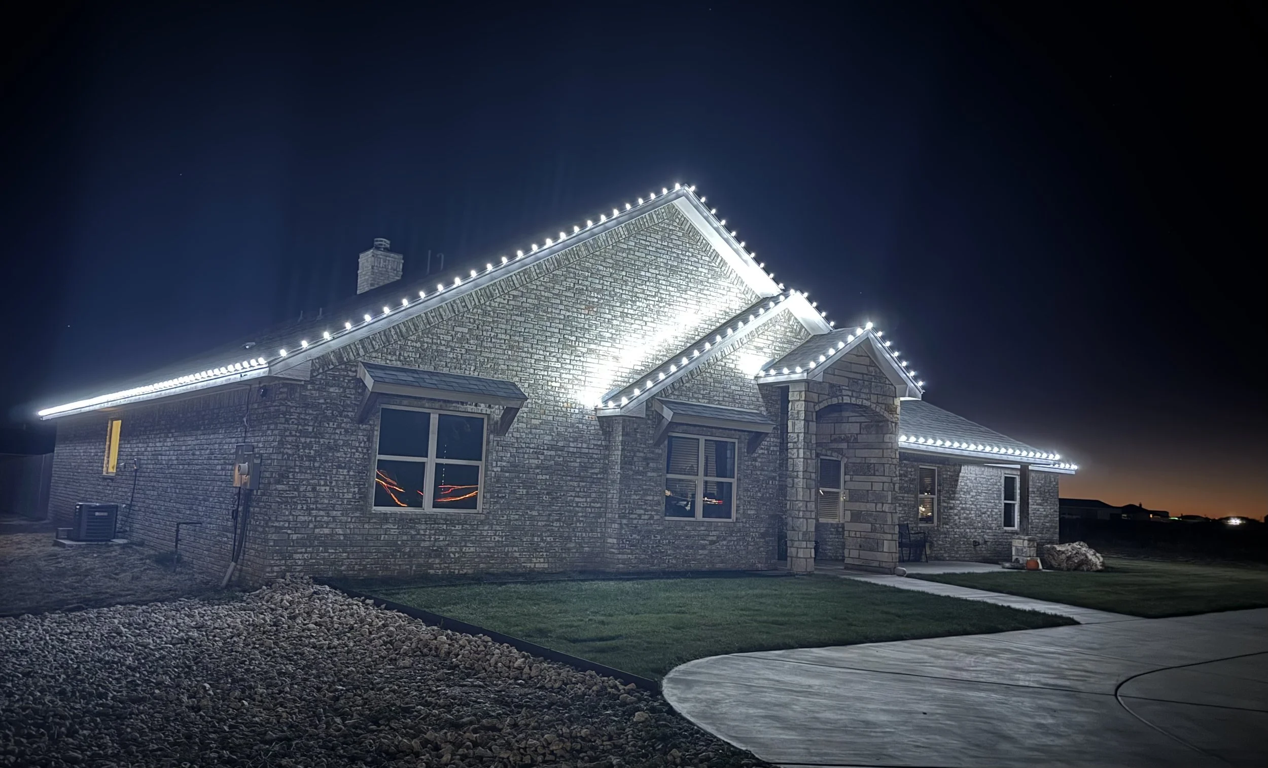 The house is decorated with white Christmas lights along the roofline, visible at night. The house is made of brick and has multiple windows. The front yard features a grassy area, a curved walkway, a gravel border, and some outdoor furniture. The sky is dark, and the house is illuminated by the string lights.