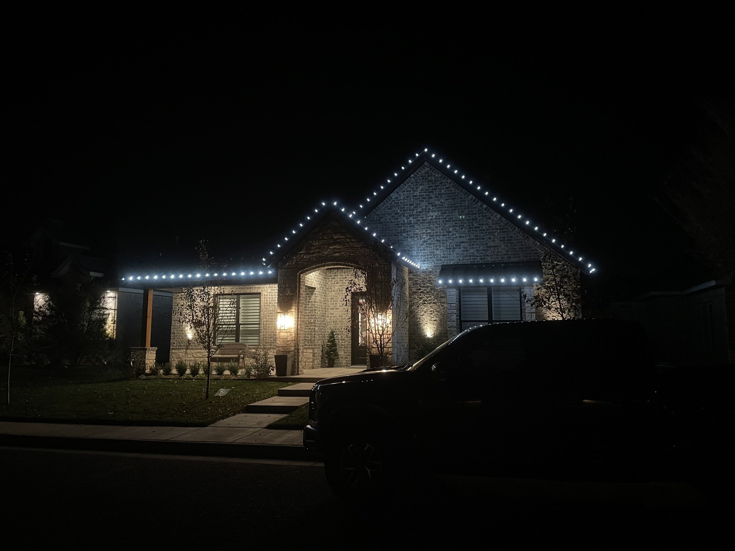Nighttime view of a house with stone and brick exterior, decorated with string lights along the roofline, with a dark sky in the background.