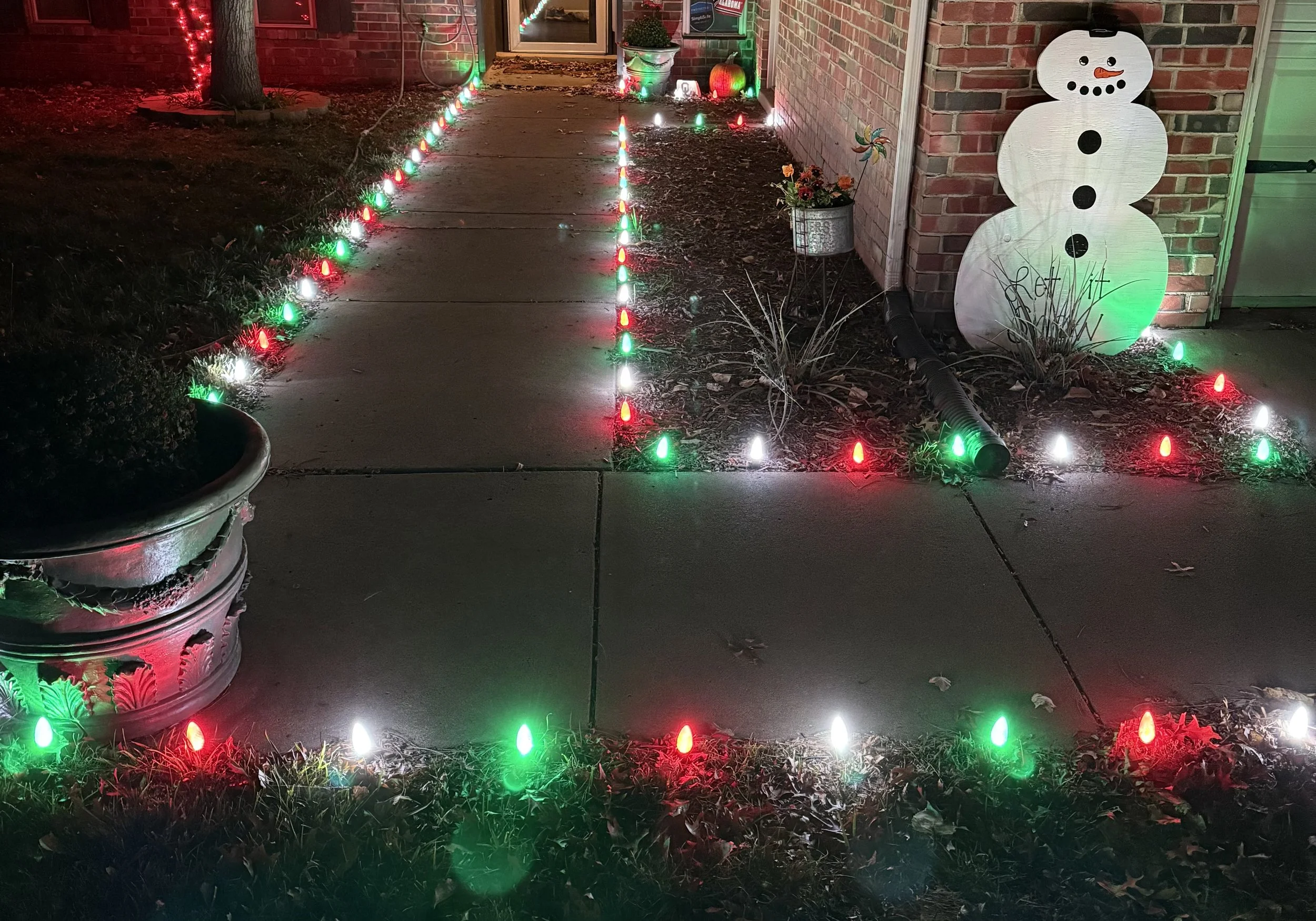 A decorated front porch at night with red, white, and green Christmas lights outlining the sidewalk and ground. There is a snowman-shaped light with a hat on the right side near the brick wall, and potted plants with flowers, a pinwheel, and an illuminated pumpkin near the door.