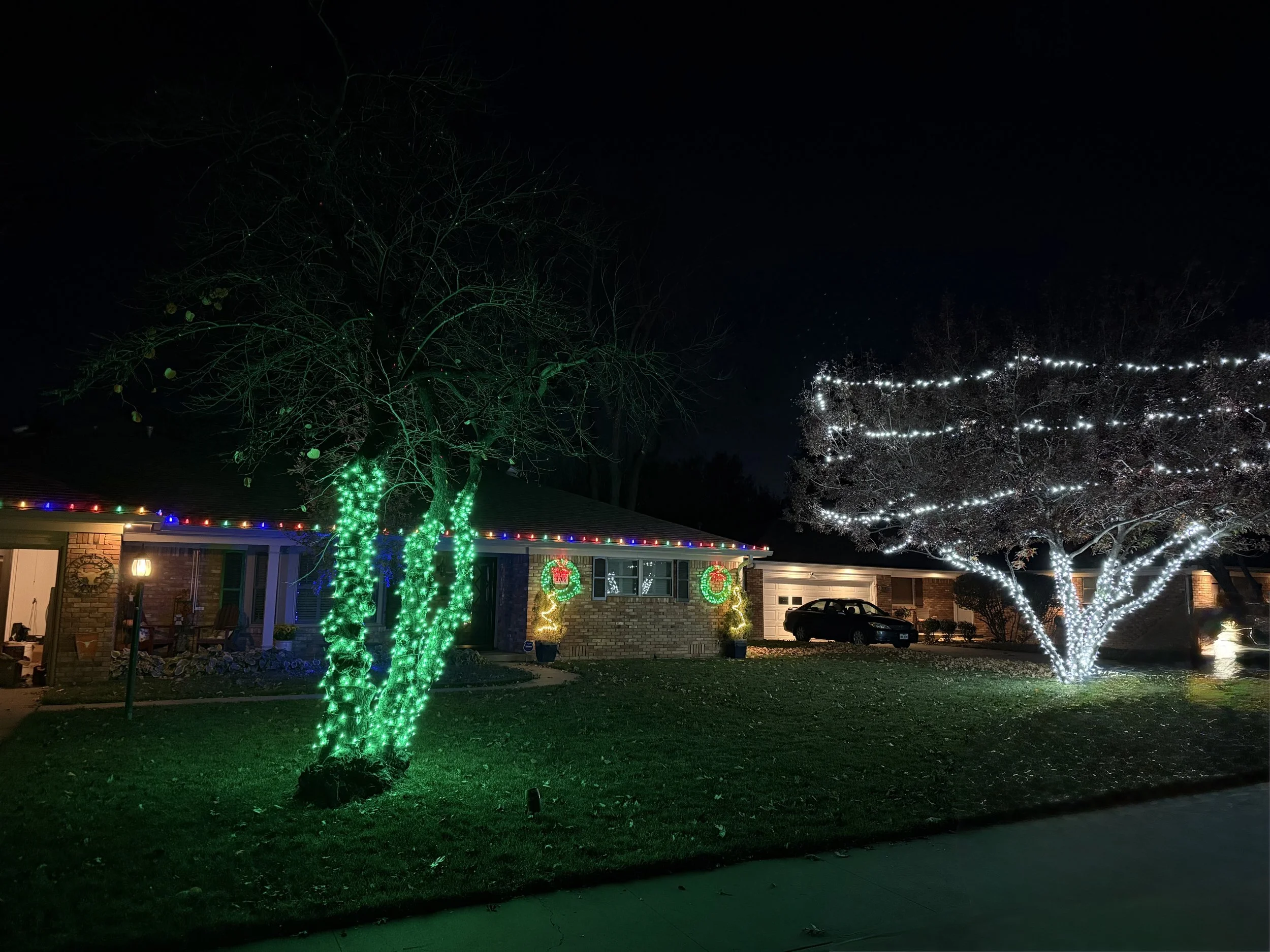 A house decorated with Christmas lights at night. There are trees with green and white string lights, and the house has multicolored lights along the roof. wreaths are visible on the house, and a car is parked in the driveway.