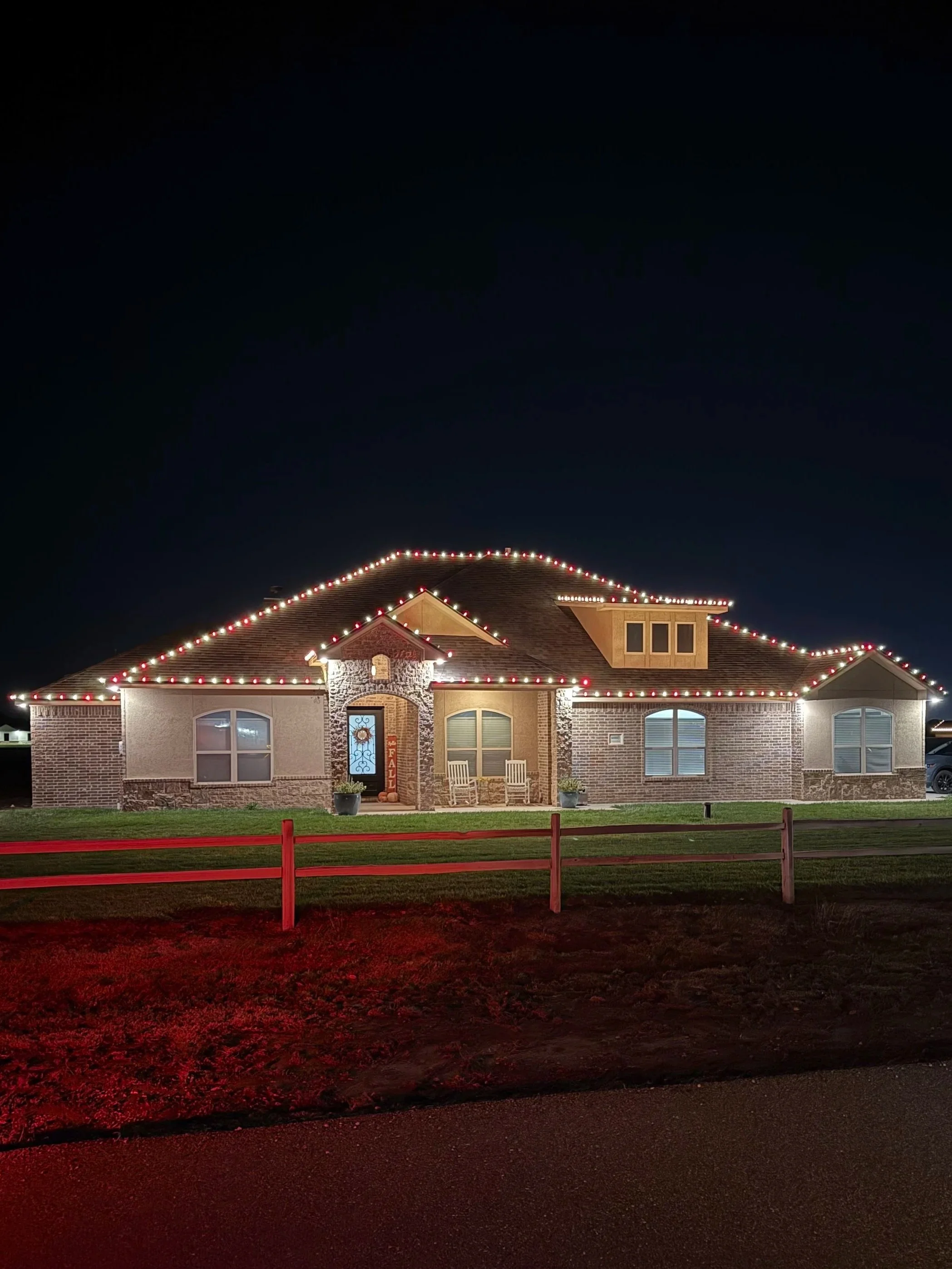 A large suburban house decorated with multicolored Christmas lights on the roofline at night, with a front yard and a white fence.