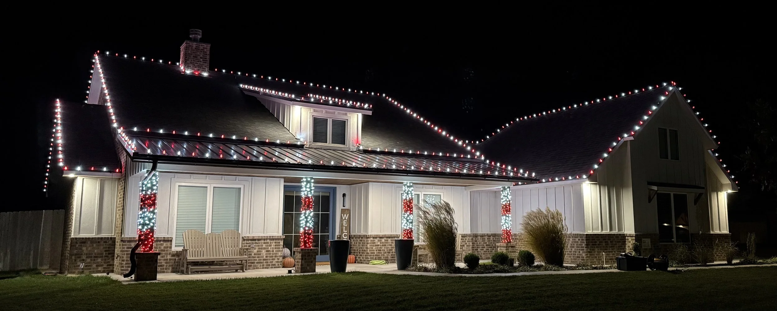 A house decorated with Christmas lights at night, including strings of red and white lights outlining the roof and columns, and lighted columns at the porch.