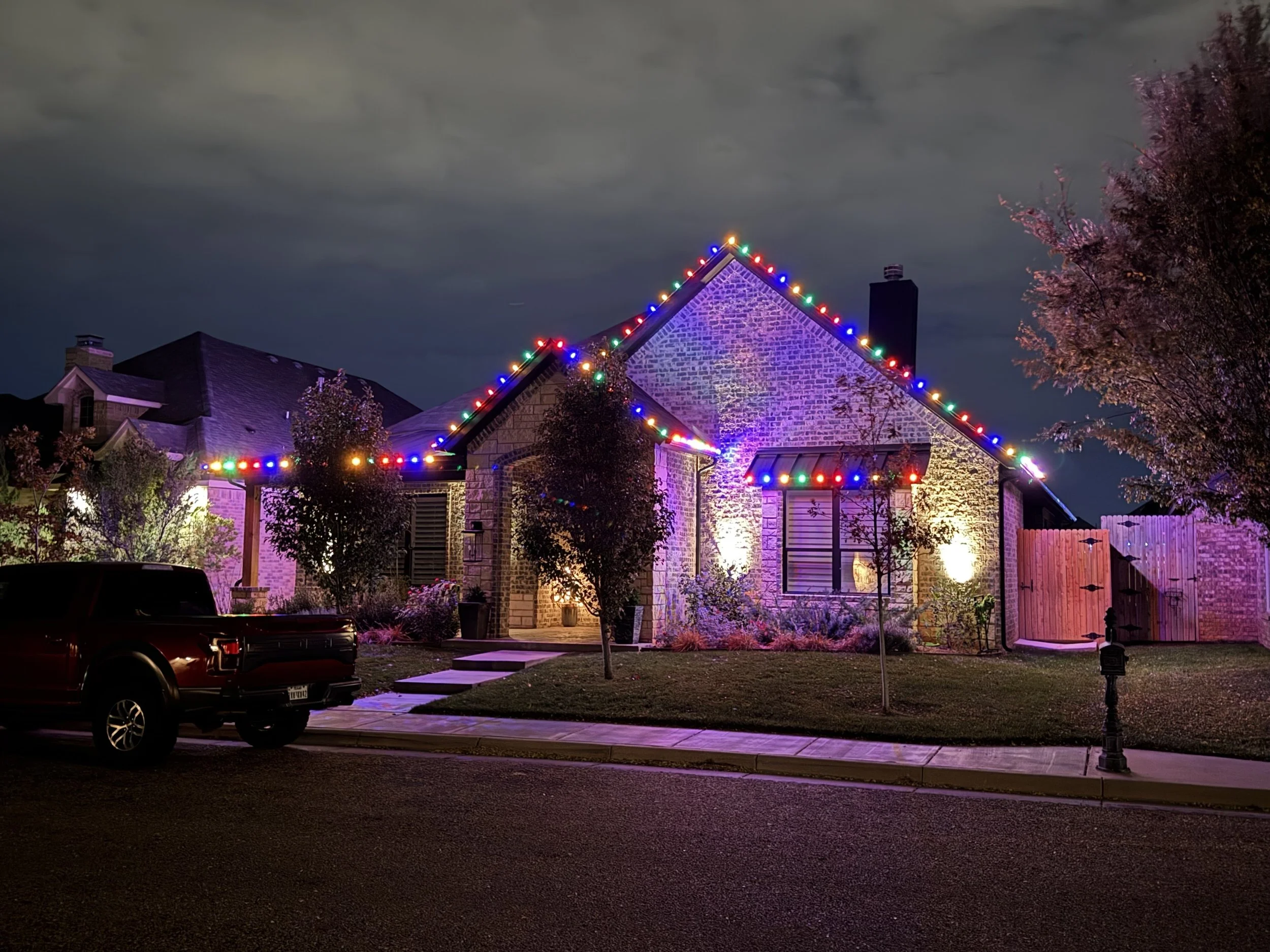 A house decorated with colorful Christmas lights at night, with trees and a sidewalk in front.