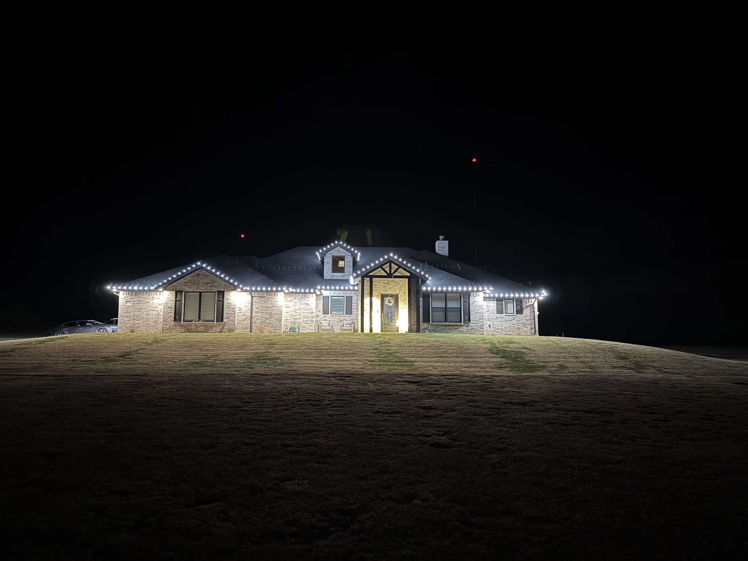 A house decorated with white string lights outlining the roof and windows, illuminated at night with a dark sky and a few distant light reflections in the background.