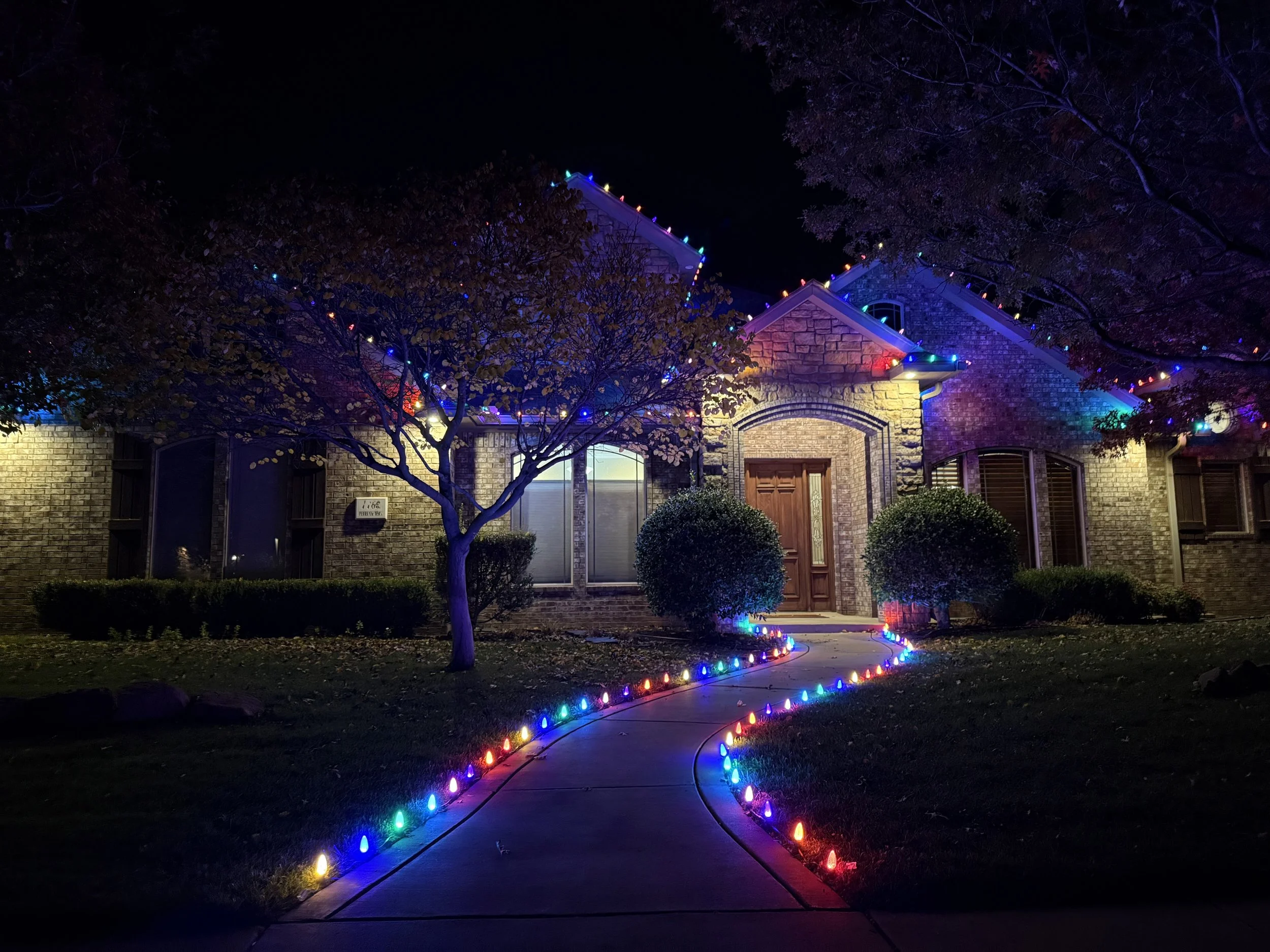 A house decorated with colorful Christmas lights along the roofline and border the driveway at night.