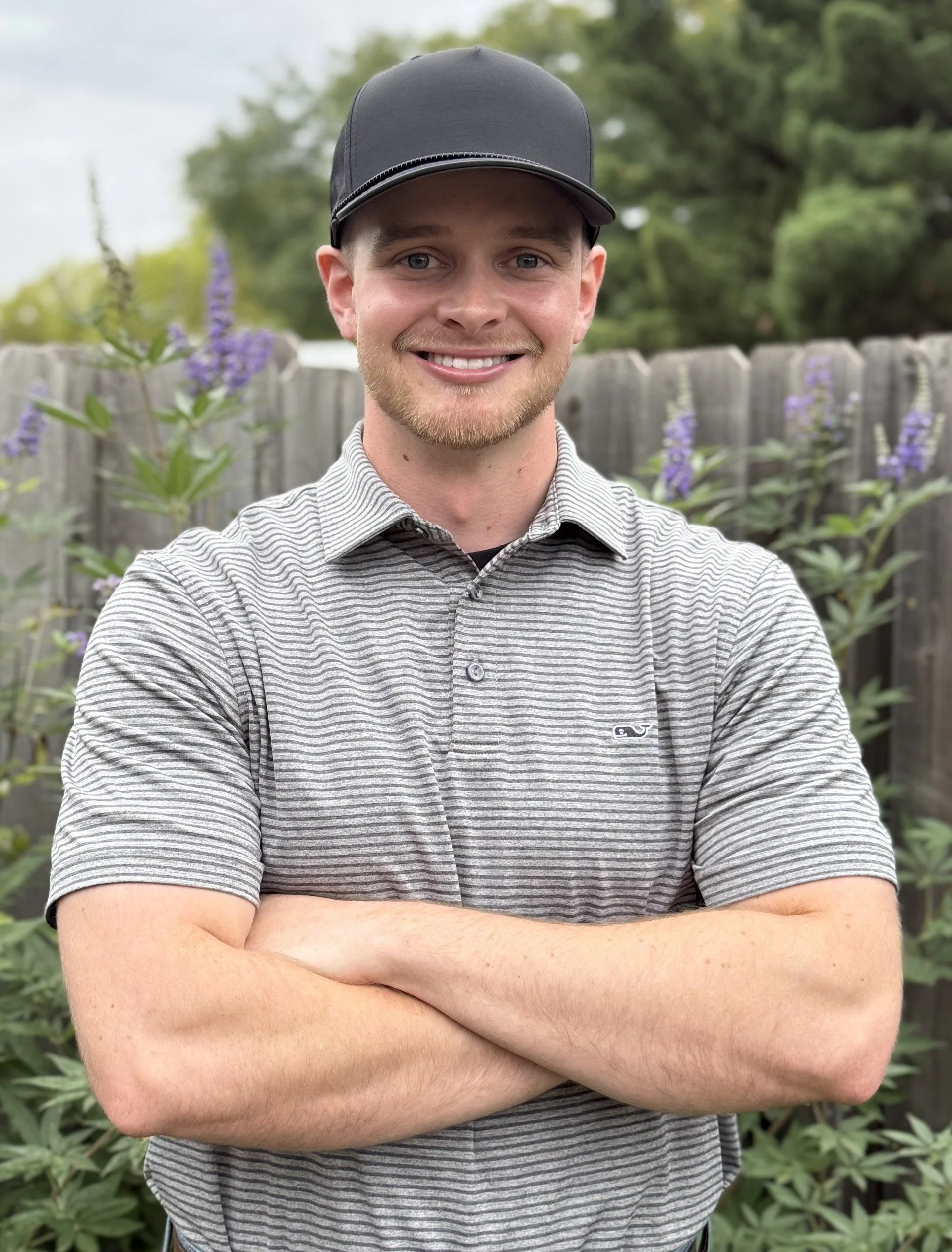 A young man with a beard and blue eyes smiling, wearing a black cap and a gray striped polo shirt, standing outdoors in front of a wooden fence with green foliage and purple flowers.