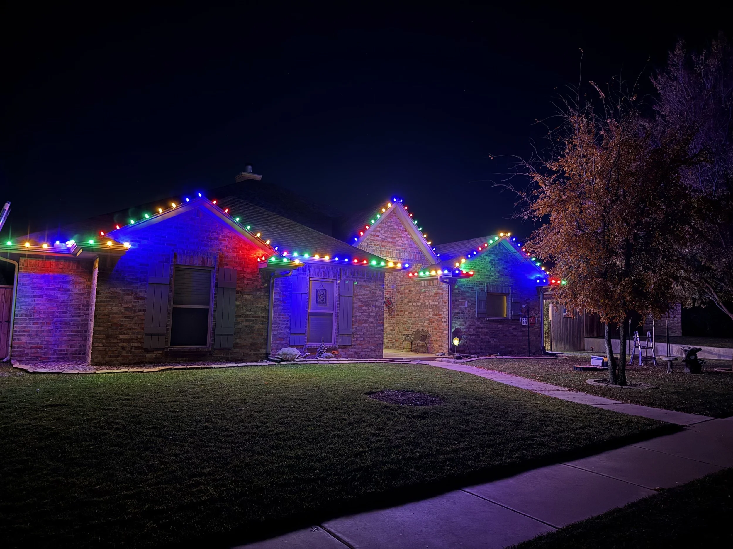 A house decorated with colorful Christmas lights on the roof, with a tree on the right side and a sidewalk in the foreground at night.