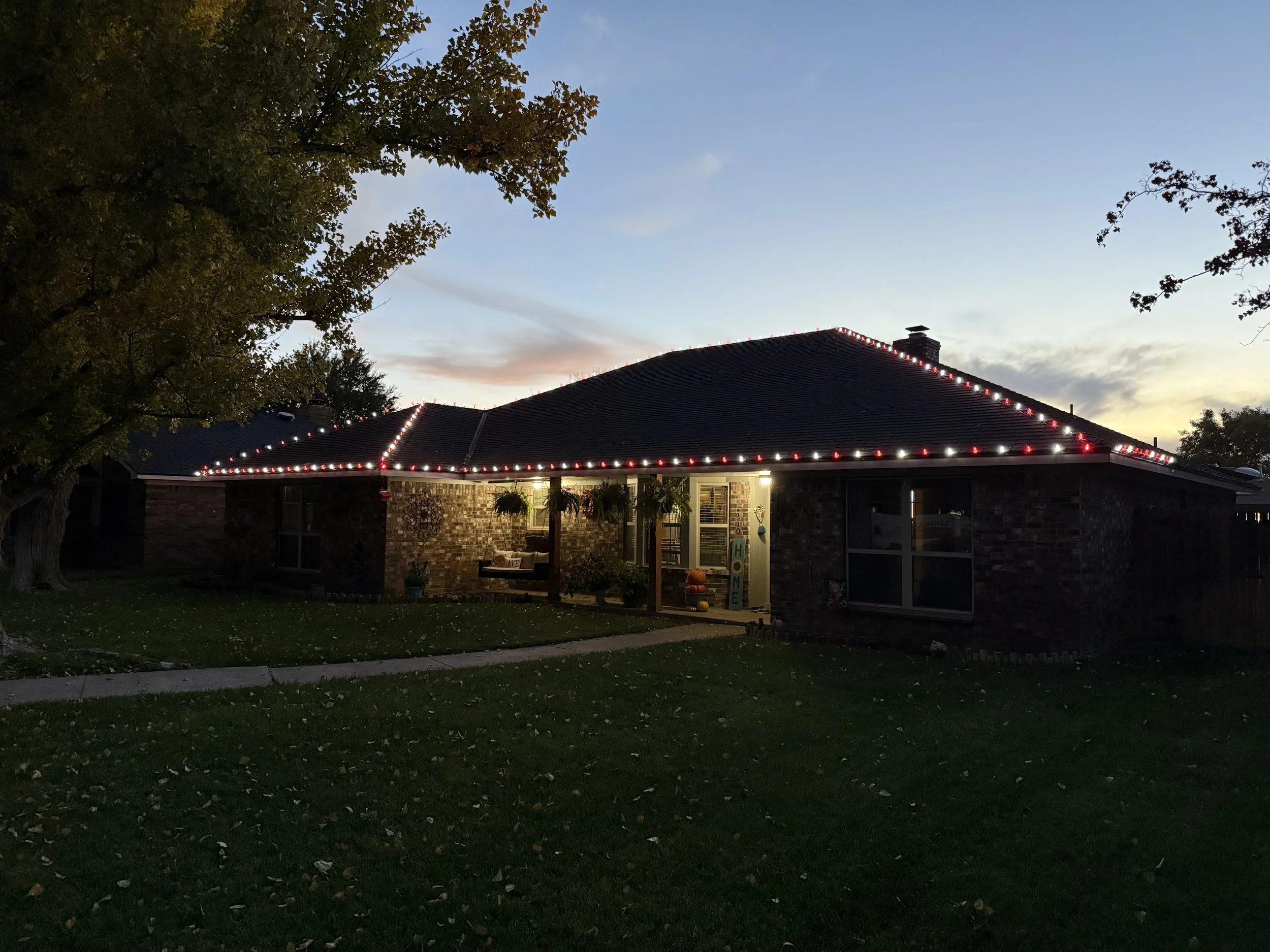 A brick house with a dark roof decorated with red and white Christmas lights, visible from the backyard at dusk, with a tree with green and brown leaves on the left and an open porch area with hanging plants and pumpkins.