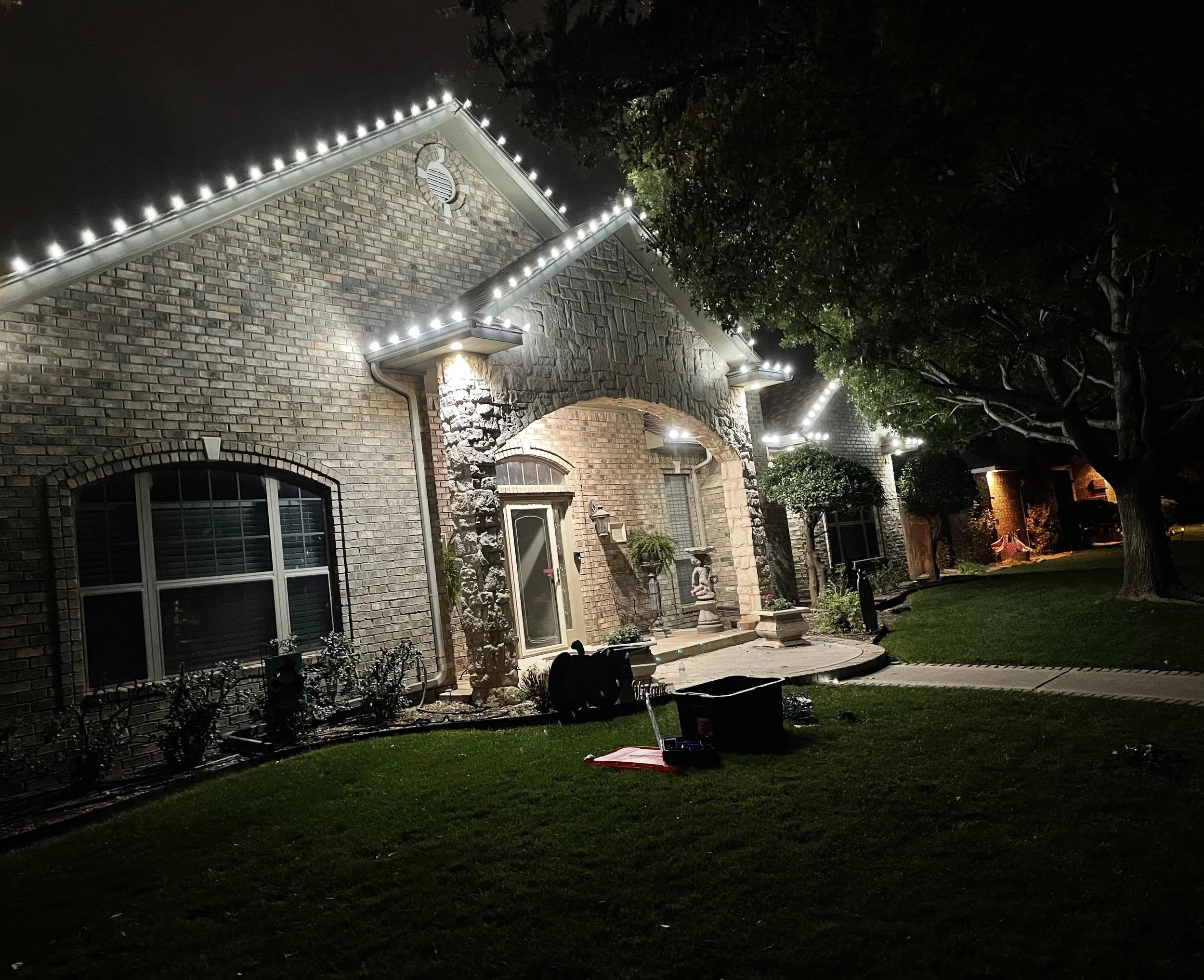 Night view of a residential house with brick walls decorated with white lights along the roofline, a garden with shrubs and trees, and home decor items near the entrance.