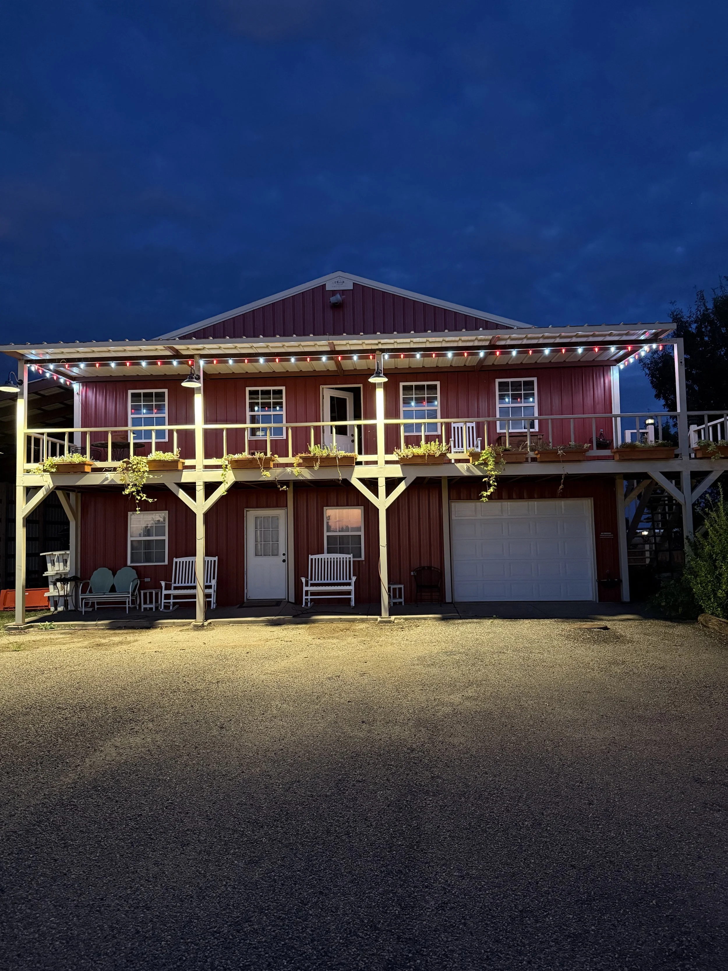 Two-story red barn with porch decorated with string lights, potted plants, benches, and outdoor furniture, under dark evening sky.