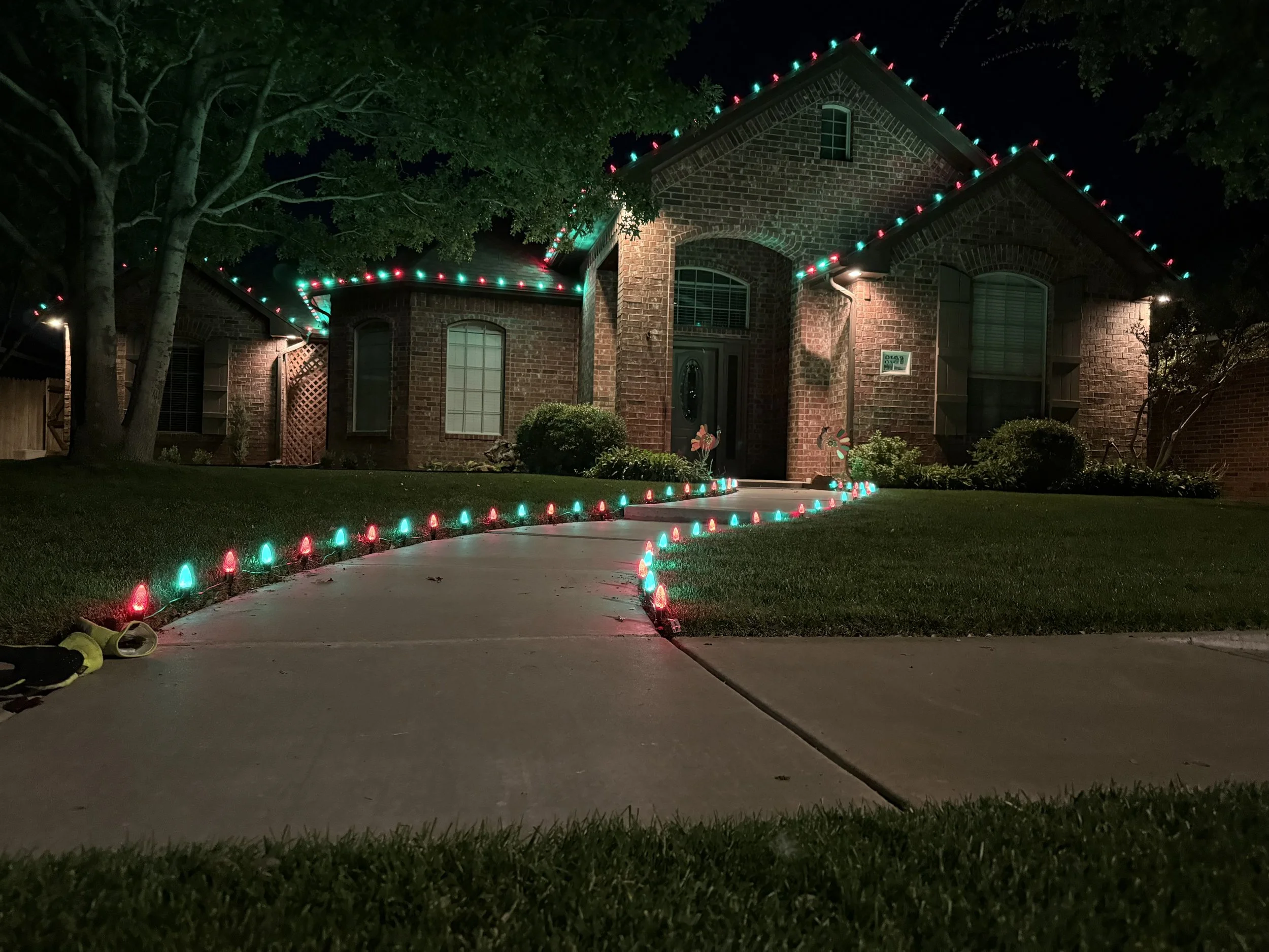 A brick house with green and red Christmas lights outlining the roof's edges at night. The front yard has a sidewalk, some bushes, trees, and silhouettes of garden ornaments, with shoes on the driveway.