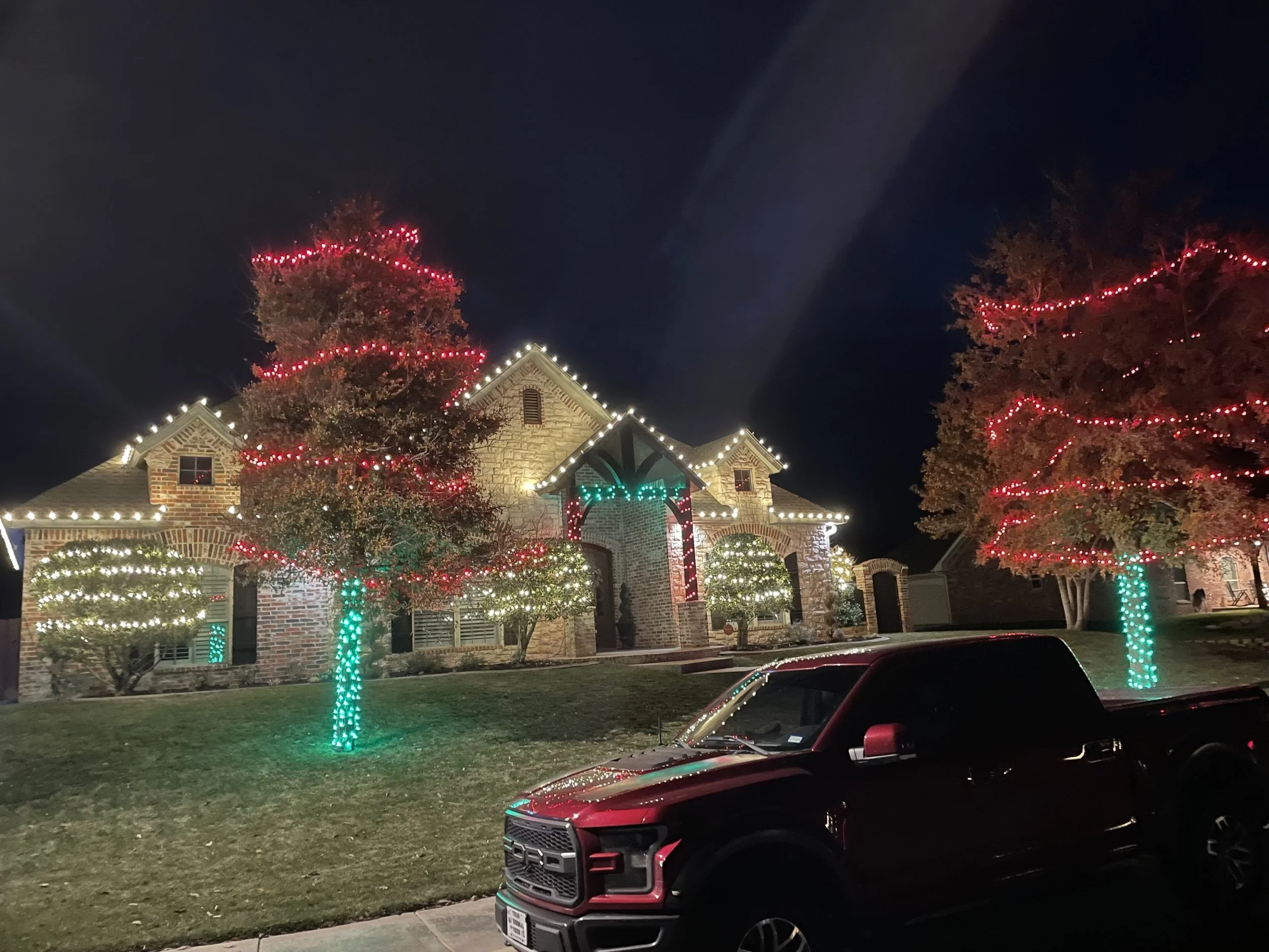Nighttime view of a house decorated with Christmas lights, including trees wrapped with green and red lights, and lights outlining the roof. A car is parked in the driveway, partially visible in the foreground.