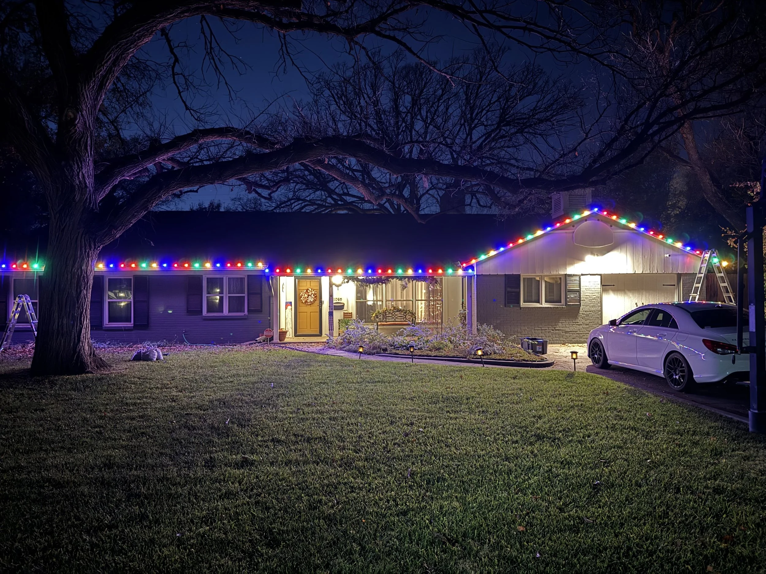 Nighttime scene of a house decorated with colorful Christmas lights along the roofline and a large tree without leaves in the yard.