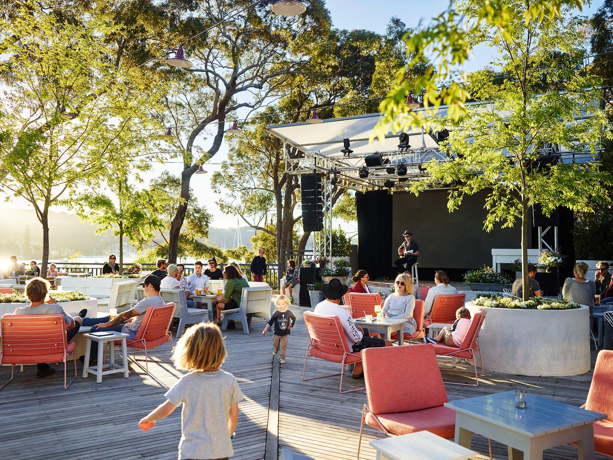 An outdoor event space with a stage, surrounded by people sitting at tables and chairs. There are trees and string lights overhead, with some sunlight filtering through the leaves. A person is performing on the stage while others are socializing or walking around.
