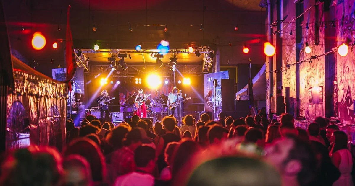 A live concert in a dimly lit indoor venue with a band playing on stage, audience watching, colorful stage lights, and string lights hanging overhead.