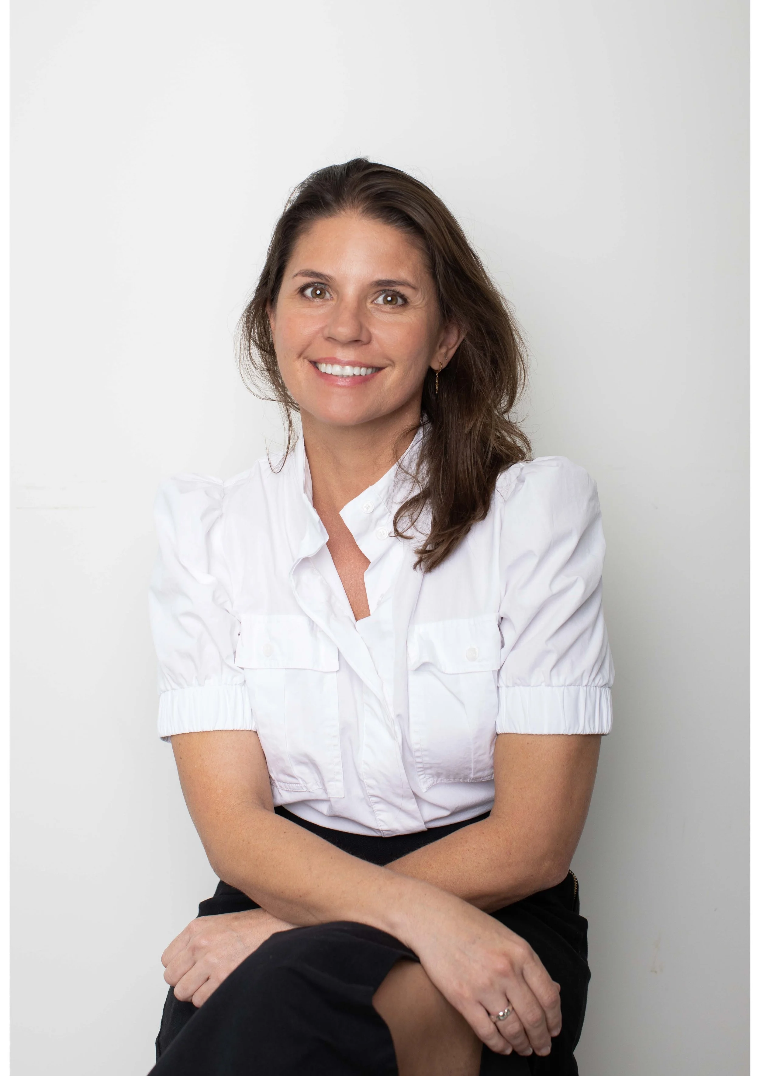 Portrait of a woman with brown hair, wearing a white blouse with puffed sleeves, sitting against a plain white background, smiling and looking at the camera.
