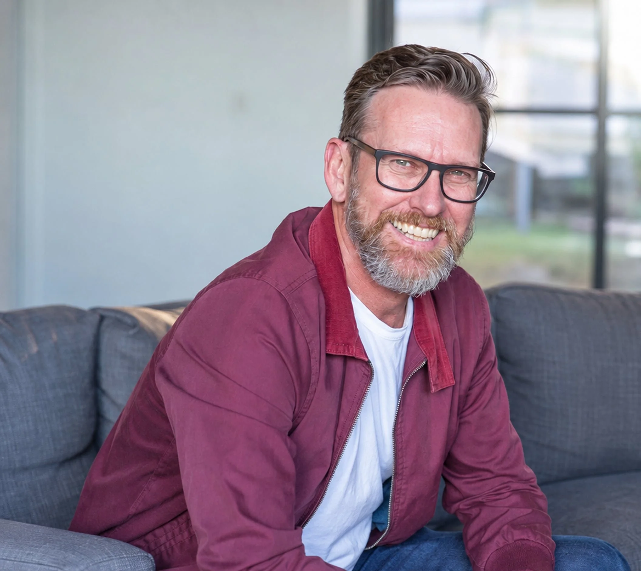 Smiling man with glasses and a beard sitting on a couch with a bright window in the background.