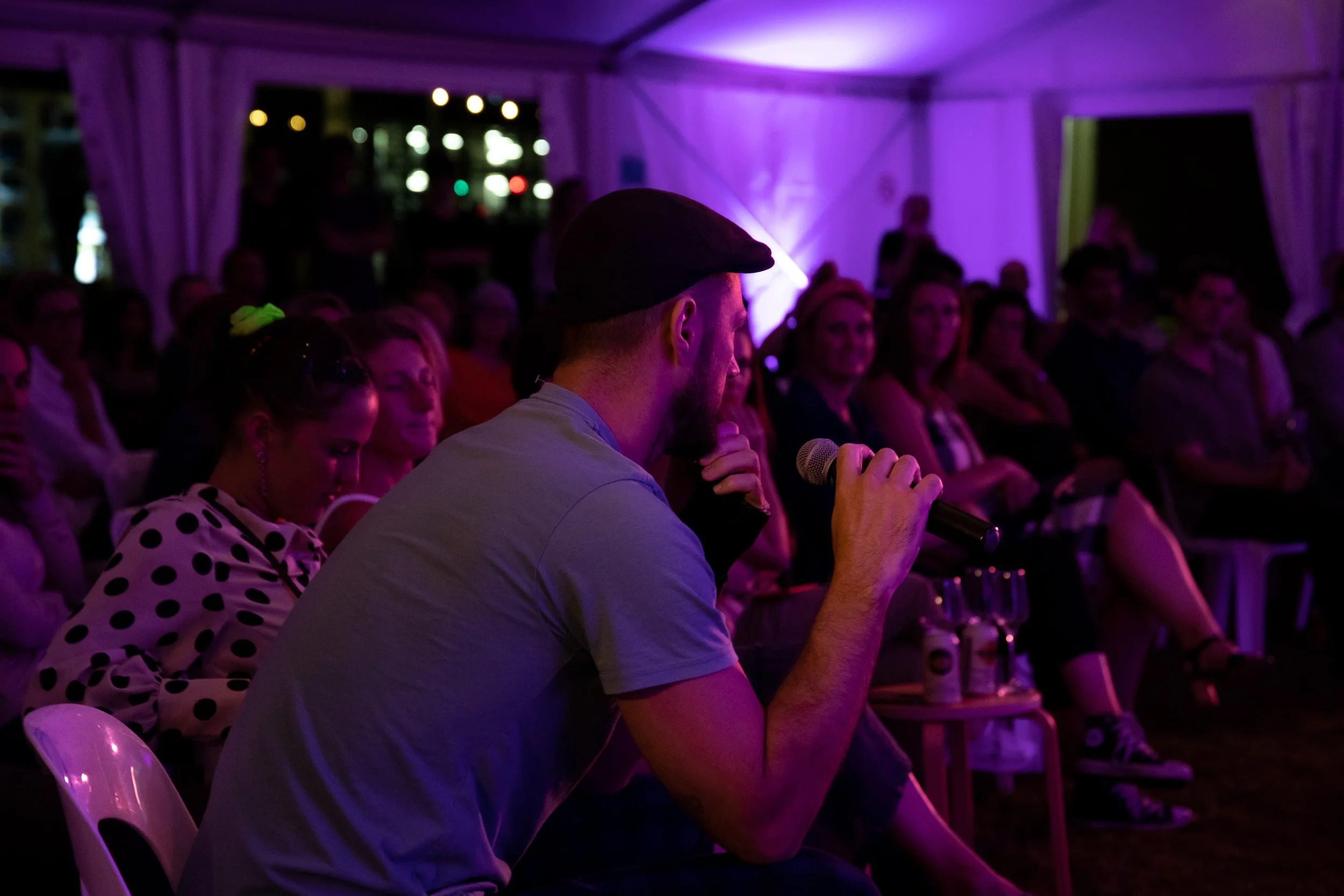 Man in a flat cap speaking into a microphone at an indoor event with an audience seated in front, purple lighting, and white curtains.