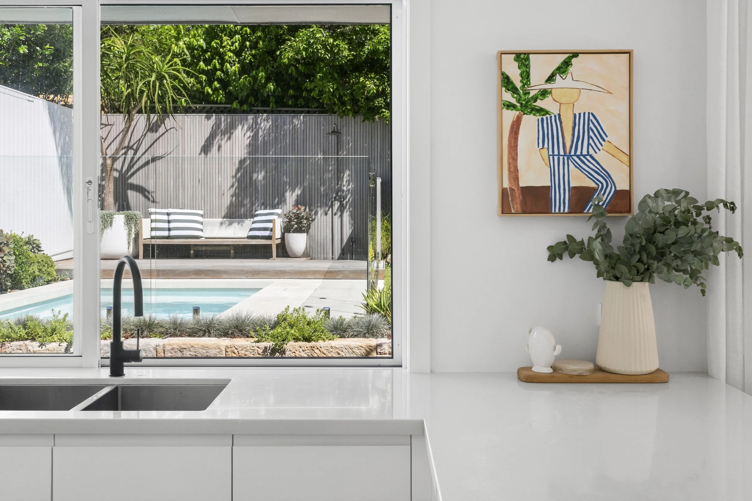 View of a backyard outdoor space through a sliding glass door from an indoor kitchen. The outdoor area features a small patio with a white bench with striped cushions, potted plants, a wooden fence, trees, and a small swimming pool with a black pool ladder.