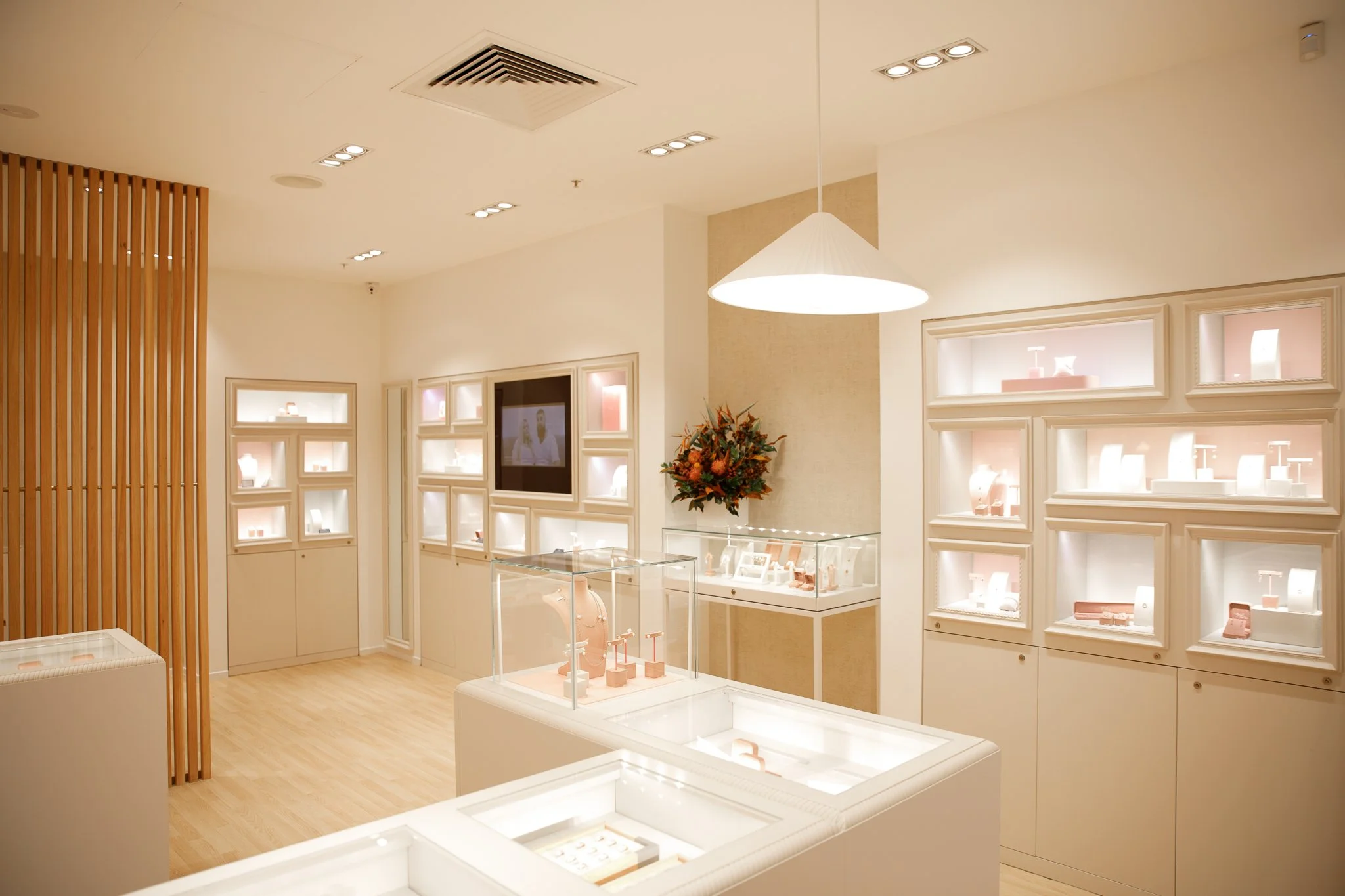 Interior of a jewelry store with white display cases and wall display shelves, a large pendant lamp, a bouquet of flowers, and a wooden slat partition.