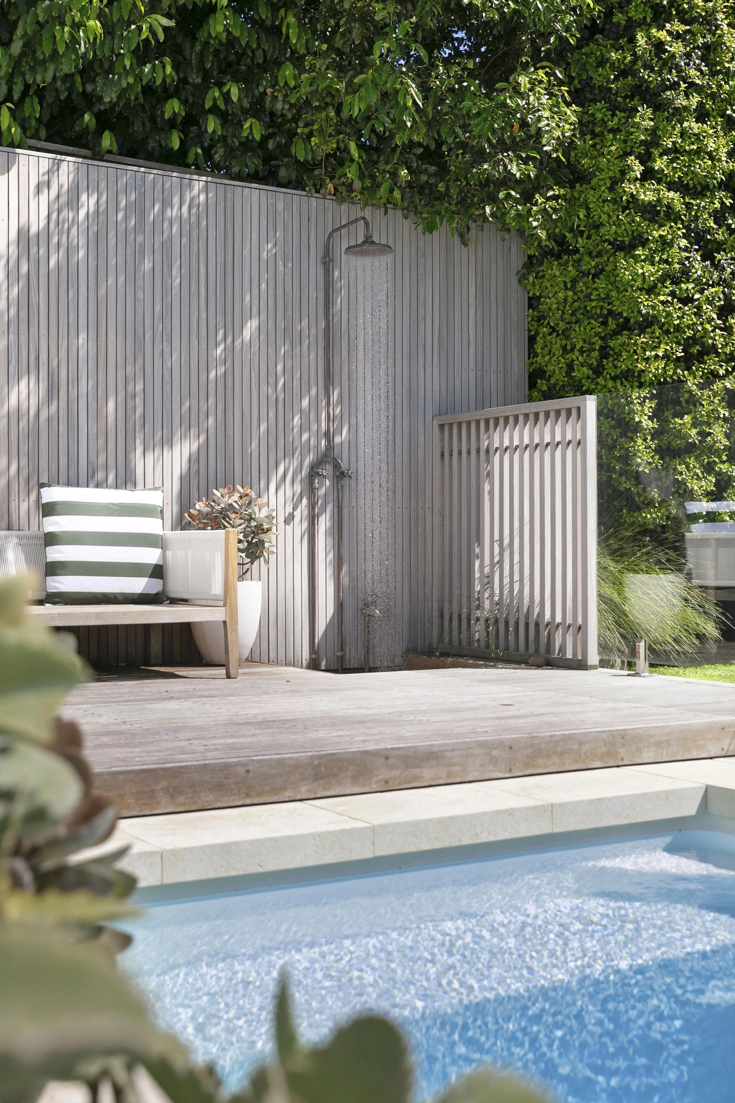 Backyard pool area with a wooden deck, a white lounge chair with green and white striped cushions, a potted plant, and an outdoor shower mounted on a wooden panel wall. The pool has blue water and there are green leafy trees in the background.
