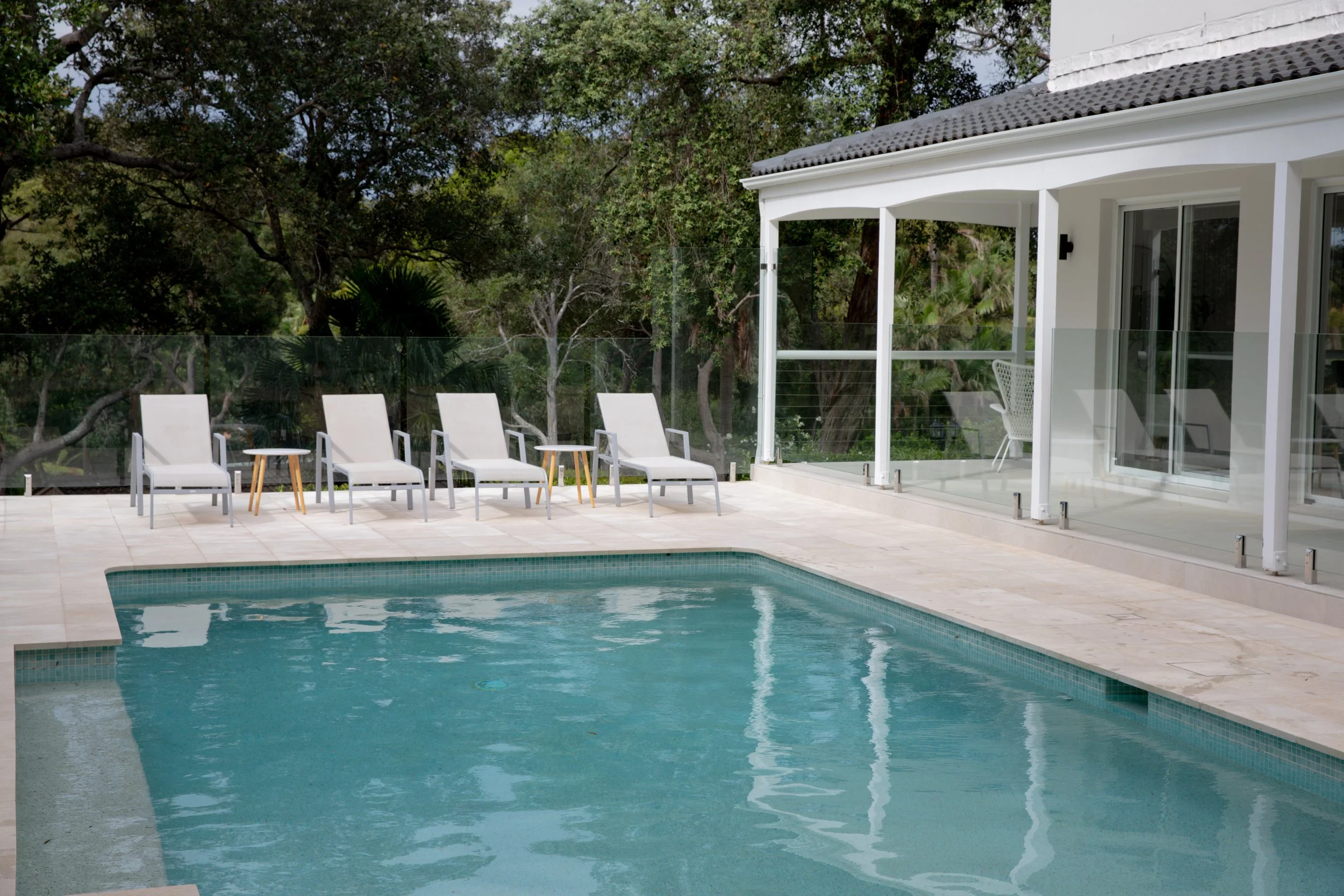 A modern backyard patio with a swimming pool, four white lounge chairs, small side tables, a glass railing, and a white house with large windows and a sliding door, surrounded by tall trees.