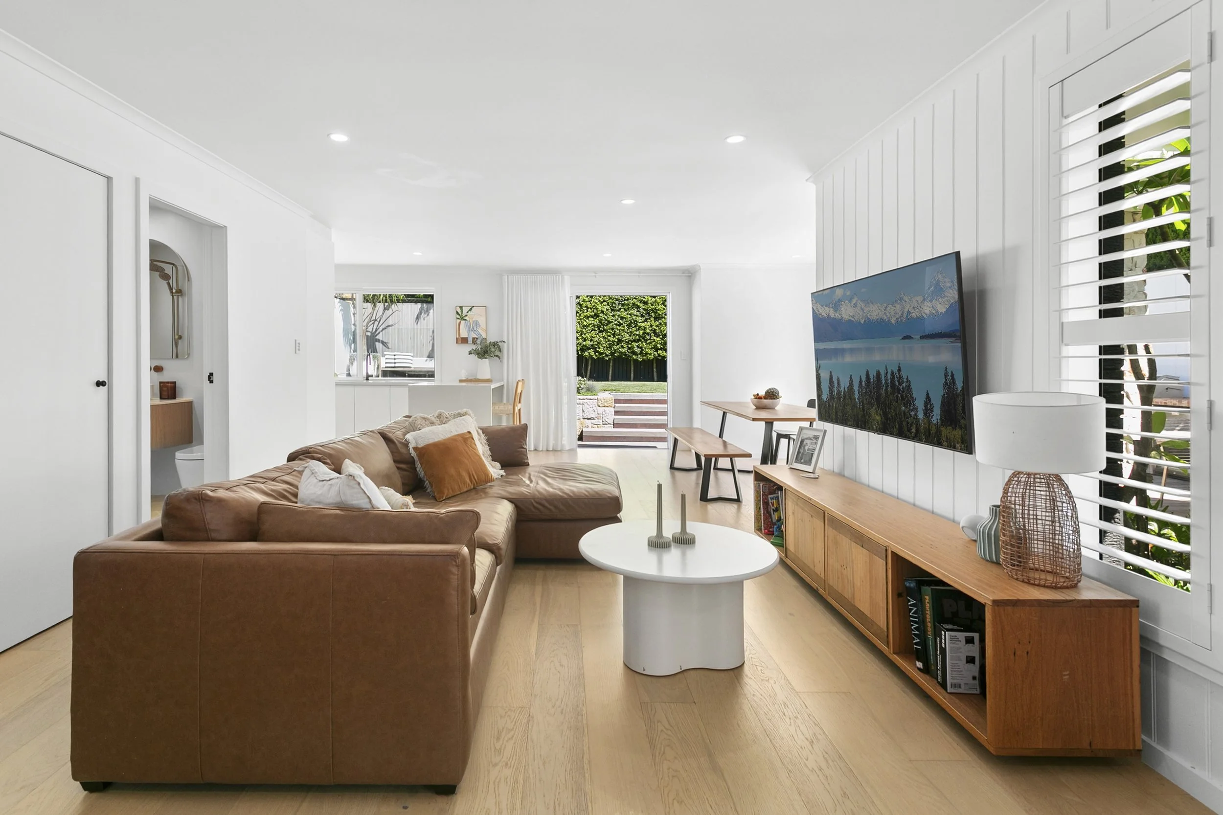 Living room with white walls, wooden flooring, a brown sofa with pillows, a round white coffee table, a wall-mounted flat-screen TV, a wooden sideboard, window with white blinds, and a view of a garden outside.