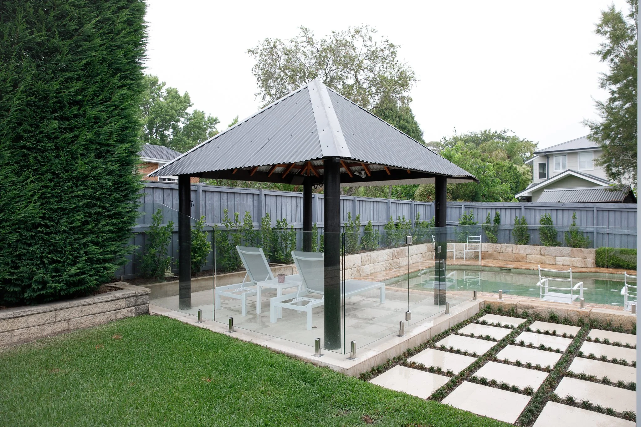 Backyard with a pool, a gazebo with a corrugated metal roof, lounge chairs, a glass fence, a stone pathway, green grass, hedges, and trees.