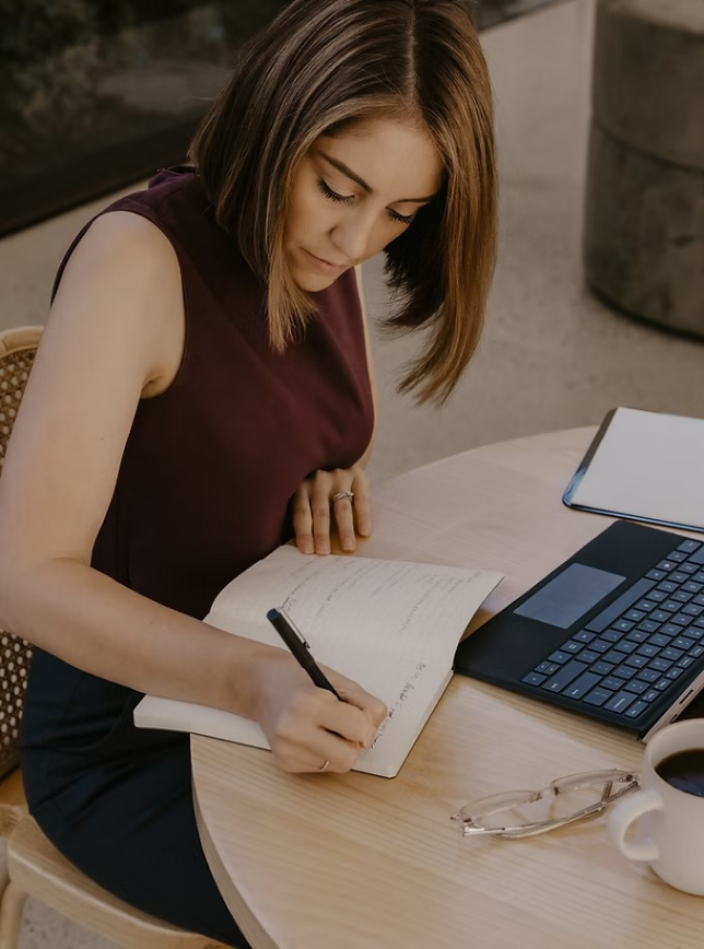 Woman sitting at a round table writing in a notebook, with a laptop, glasses, and a cup of coffee nearby.