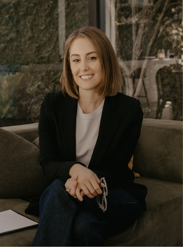 A picture of Dianne Casotti, who is a registered Psychologist with shoulder-length brown hair wearing a black blazer and white shirt, sitting on a couch with a notepad on her lap.