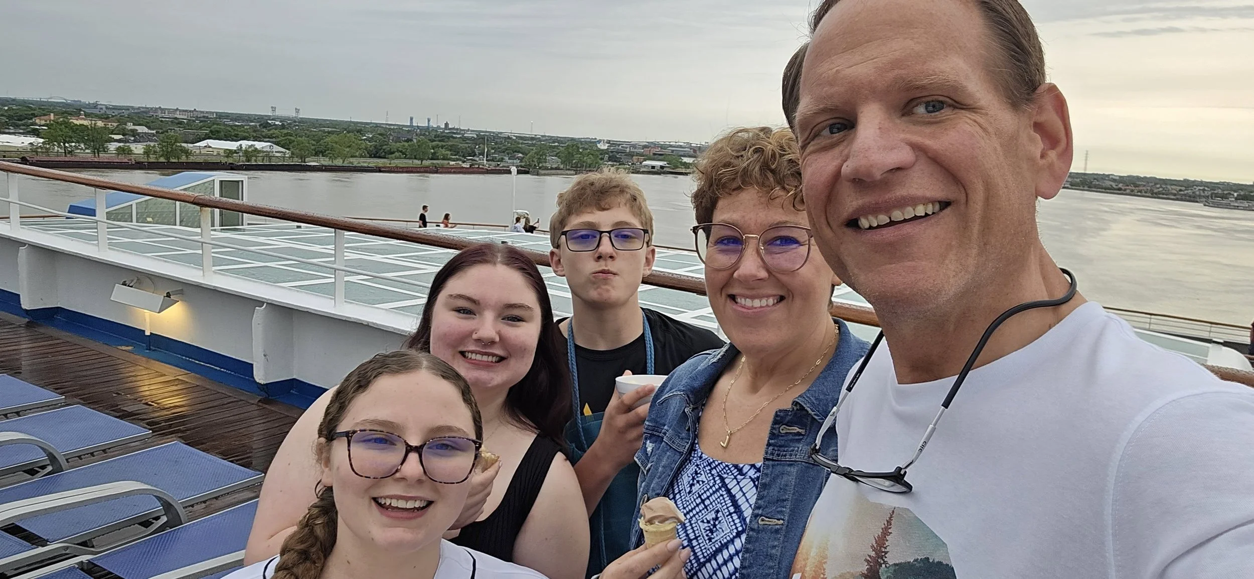 A family of five on a cruise ship deck, smiling at the camera, with a river and cityscape in the background.