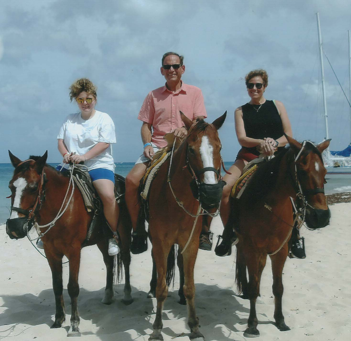 Three people riding horses on a beach, with waves and a sailboat in the background.