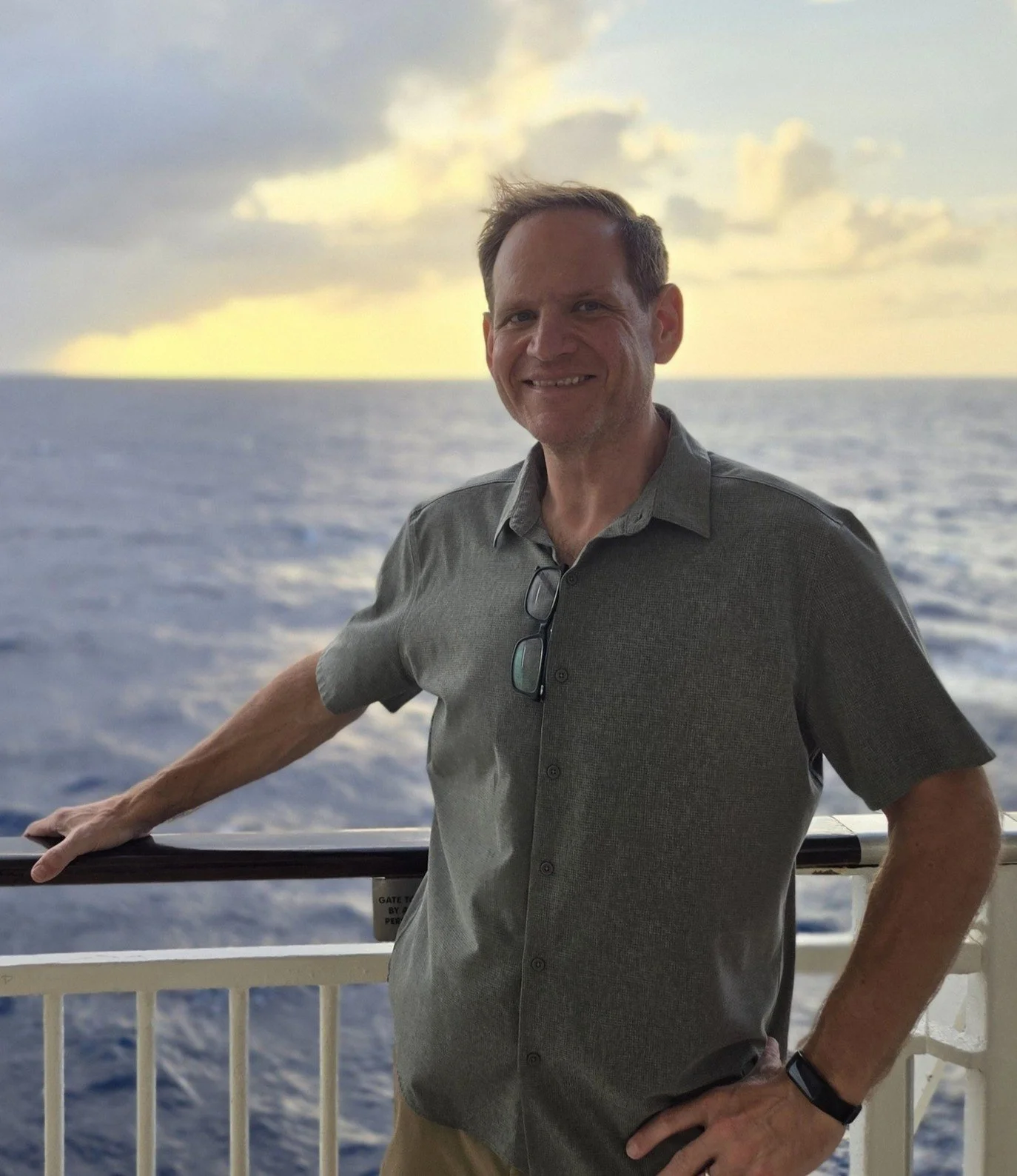 A man with short brown hair smiling, wearing a gray button-up shirt with glasses hanging on his collar, standing near a railing on a boat, with a large body of water and cloudy sky in the background.