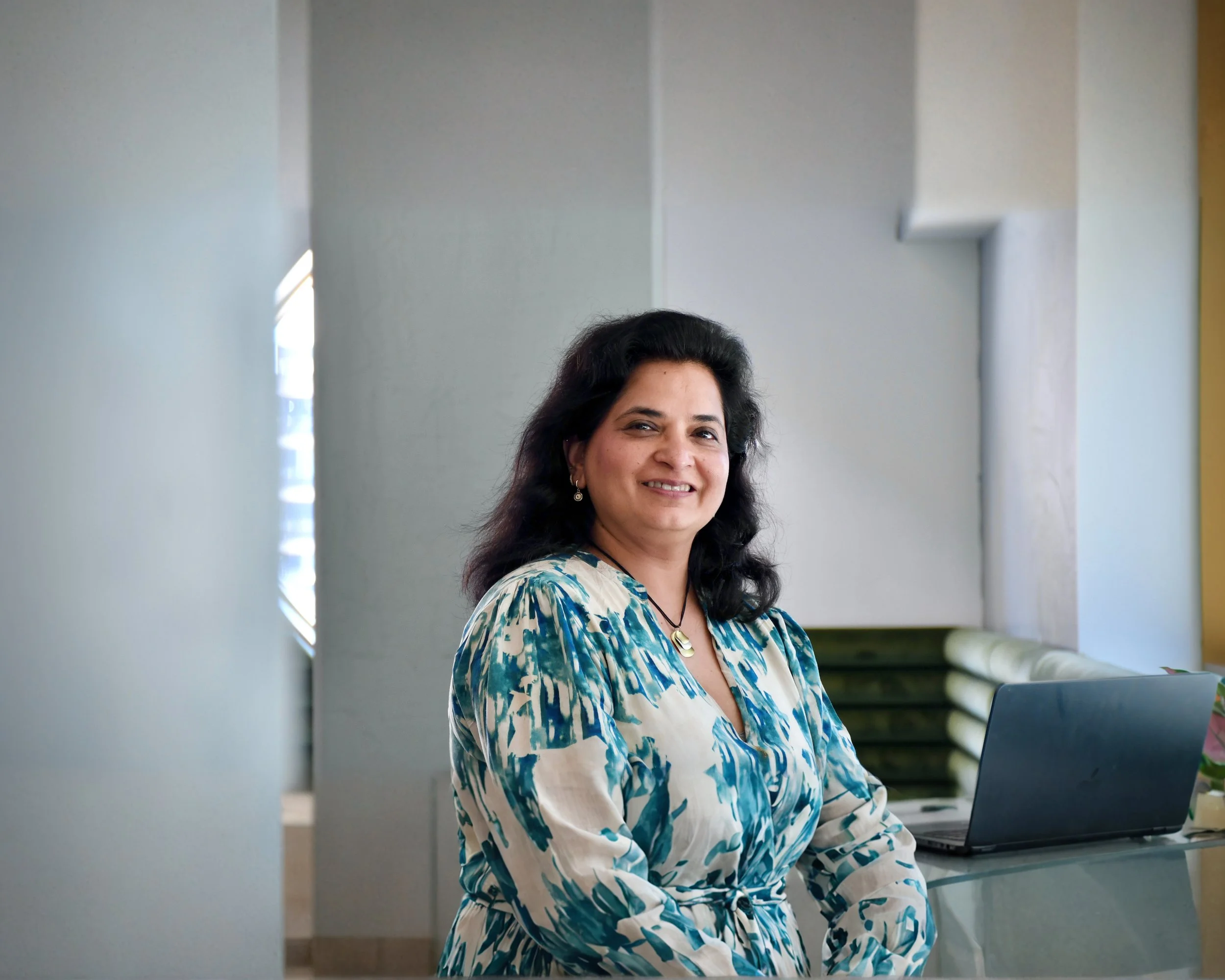 A woman with dark hair, wearing a blue and white patterned blouse, sitting at a desk with a laptop in a modern office setting.